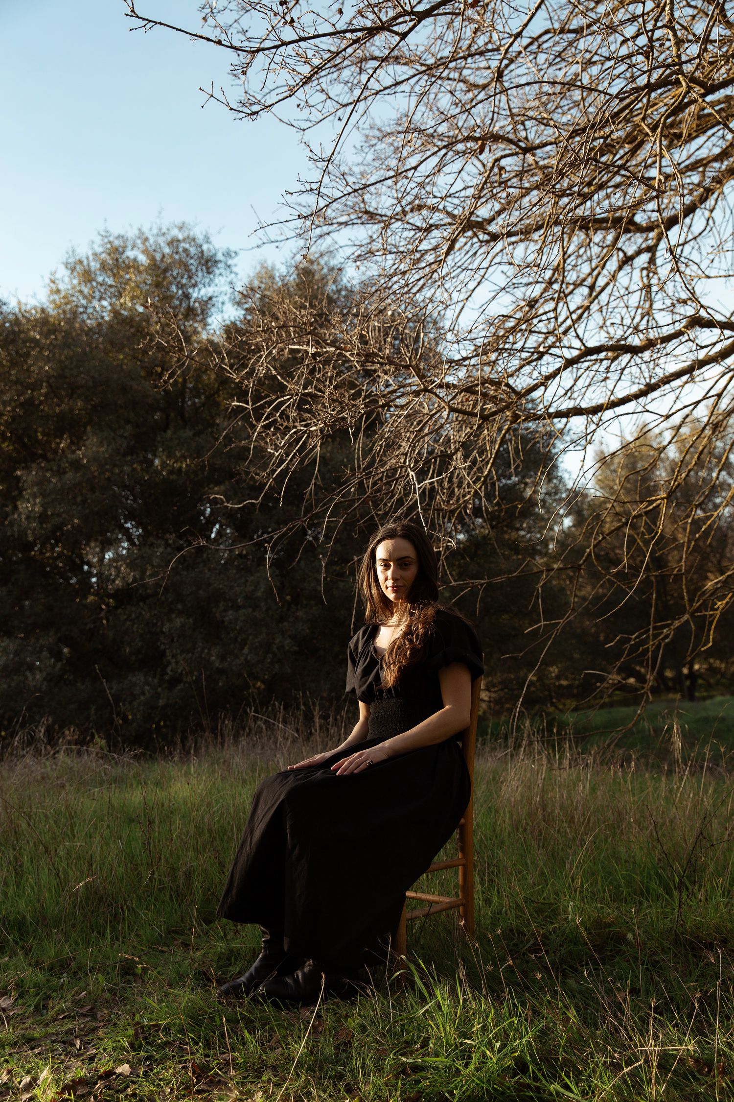 Figure in a black dress poses in a meadow under branching trees during golden hour.