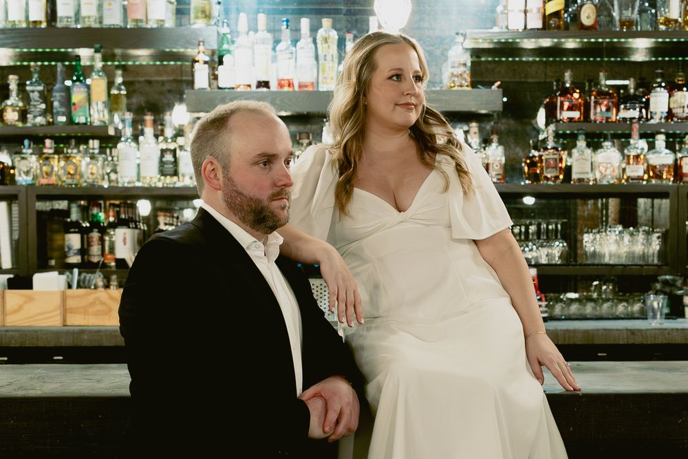 A bride and groom pose together at a well-stocked bar with glasses and bottles visible in the background.