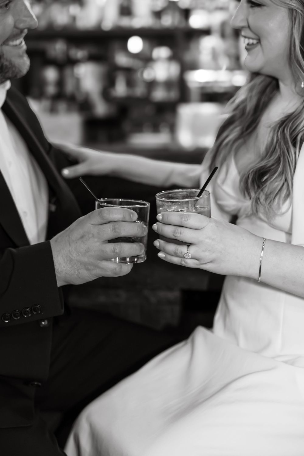 A couple in formal attire toasts with cocktail glasses at a bar in an intimate black and white moment.