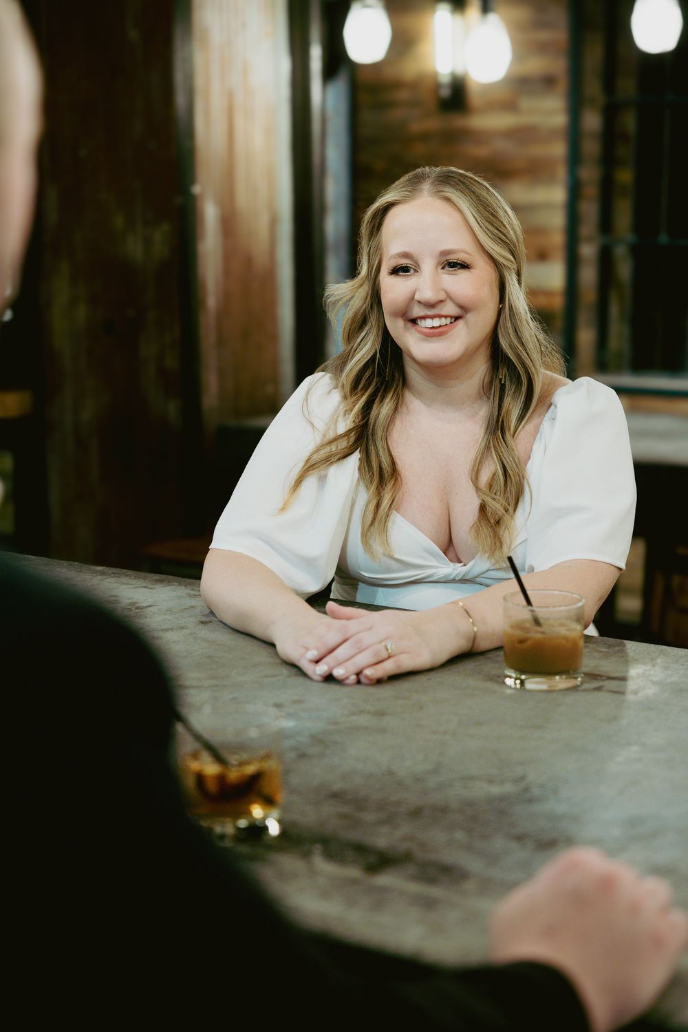 Woman in white top smiling at bar counter with cocktail drinks in dimly lit restaurant setting.