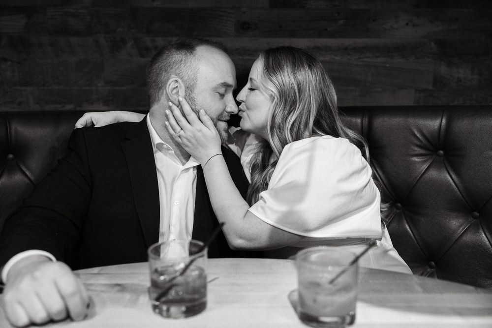Black and white romantic moment captured between couple sharing intimate kiss at restaurant booth with drinks on table.