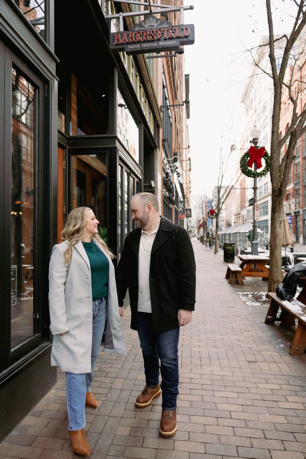 A couple walks together on a brick sidewalk past storefronts with holiday decorations during winter.