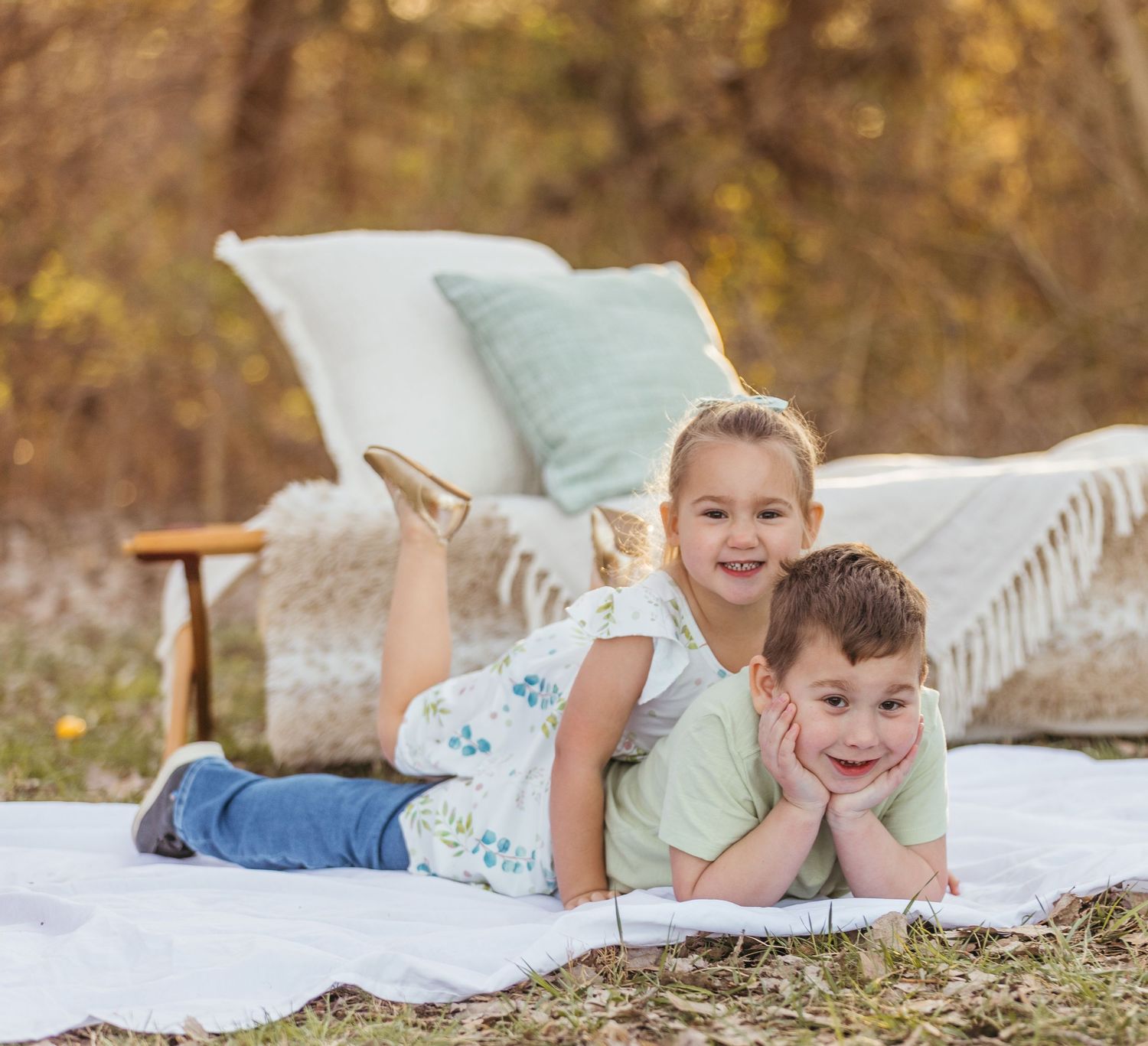 Two children laying on a white blanket with pillows outdoors in autumn scenery.