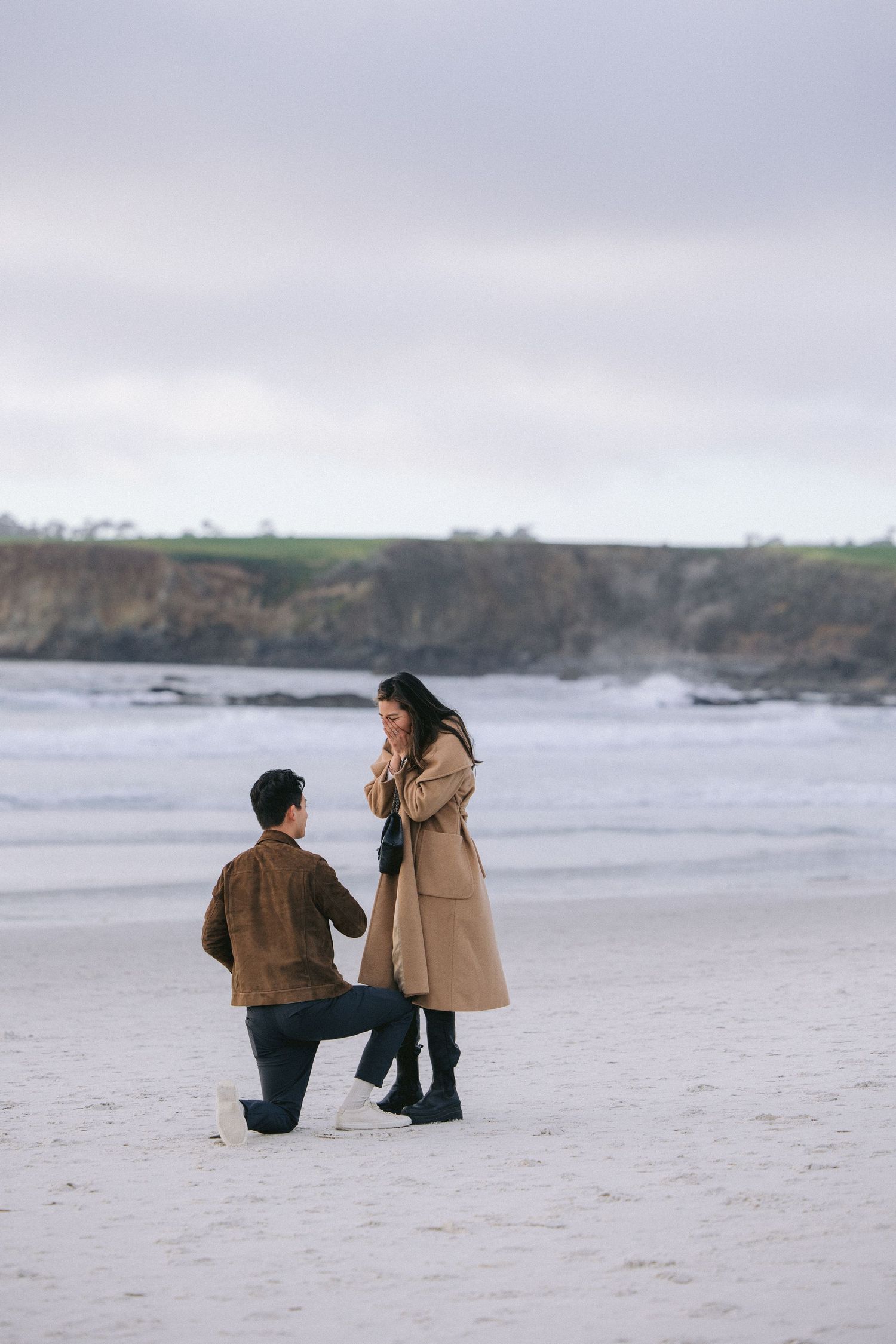 A romantic beach proposal scene with cliffs and ocean in background during an overcast day.