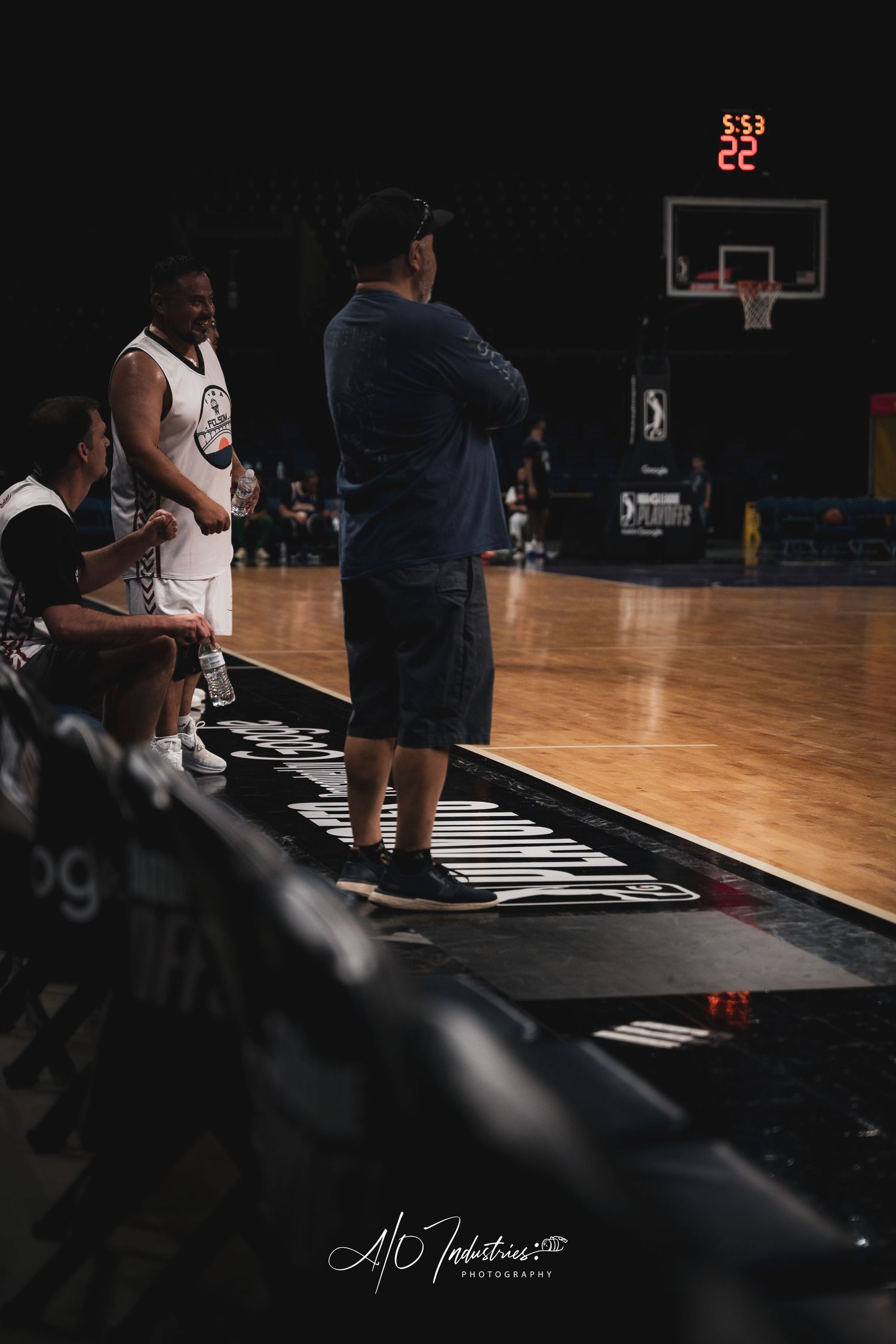 Basketball coach stands with arms crossed courtside while players rest on bench during indoor arena game.
