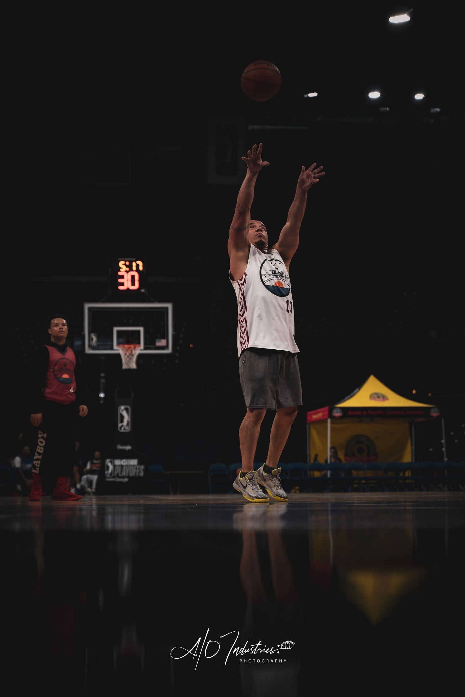 Basketball player shooting a jump shot in a dark indoor arena with scoreboard and hoop visible in background.