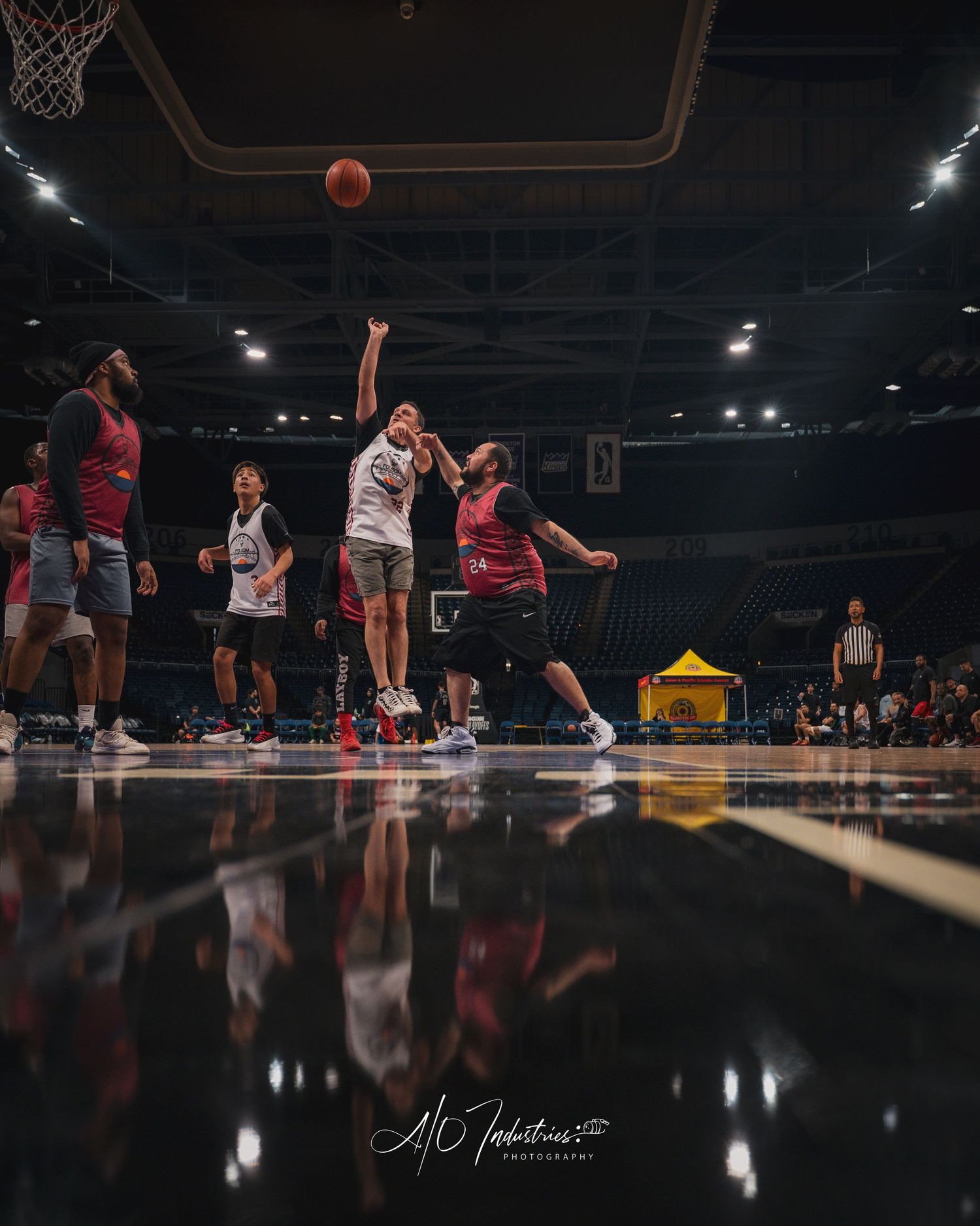 Players compete for basketball near hoop during indoor arena game with reflective court floor visible.