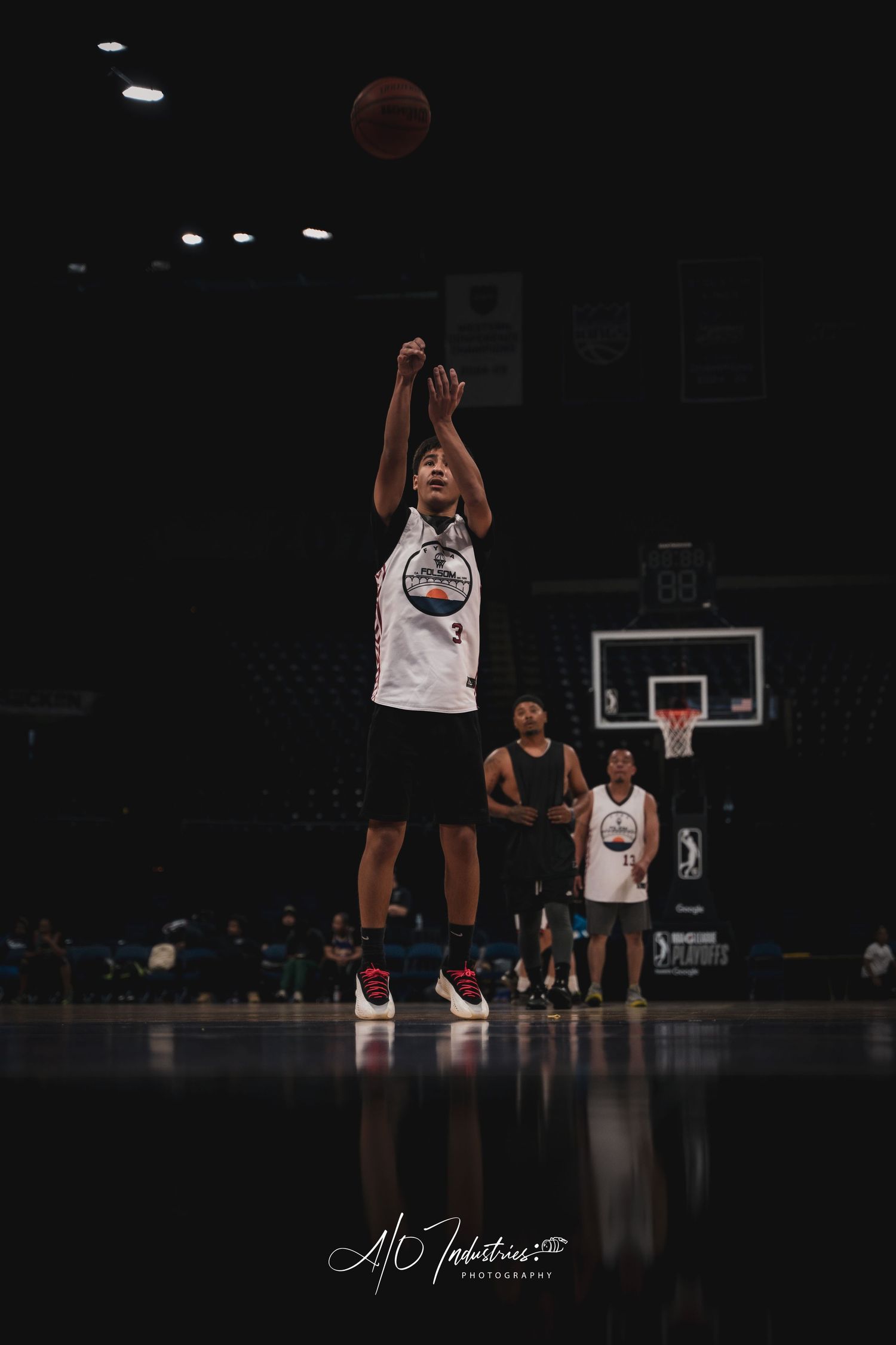 A player in a white jersey releases a jump shot in a dimly lit basketball arena.