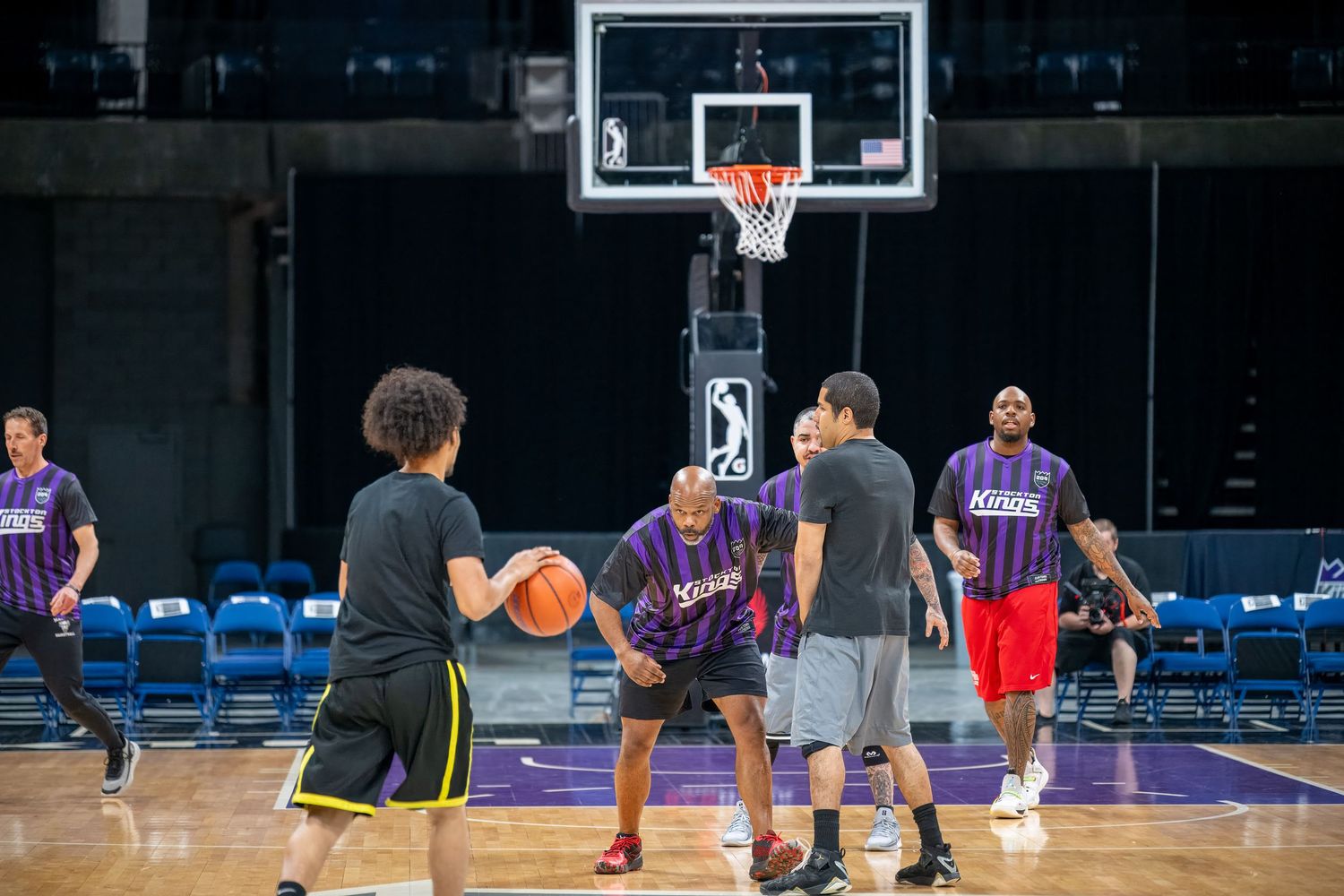 Players gather around a basketball hoop during practice on a purple indoor court.