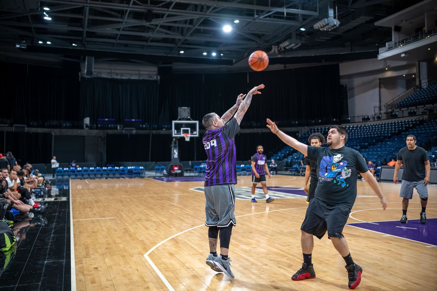 Basketball player takes a jump shot while defender attempts to block on indoor court.