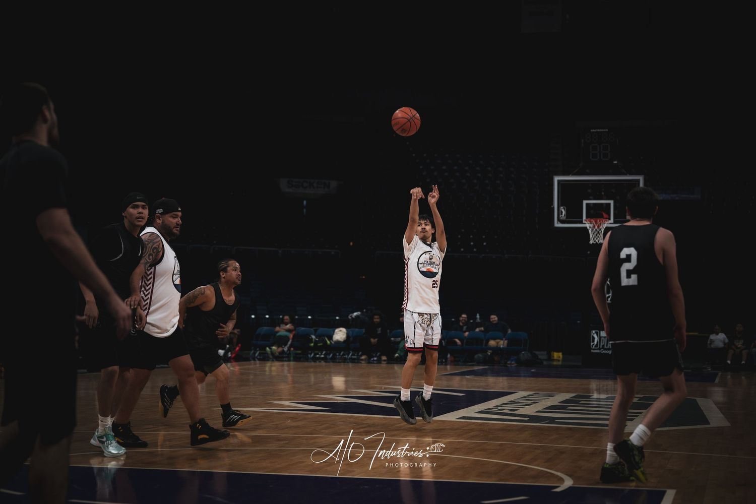 A basketball player shoots a mid-range jumper during an indoor game with defenders nearby.
