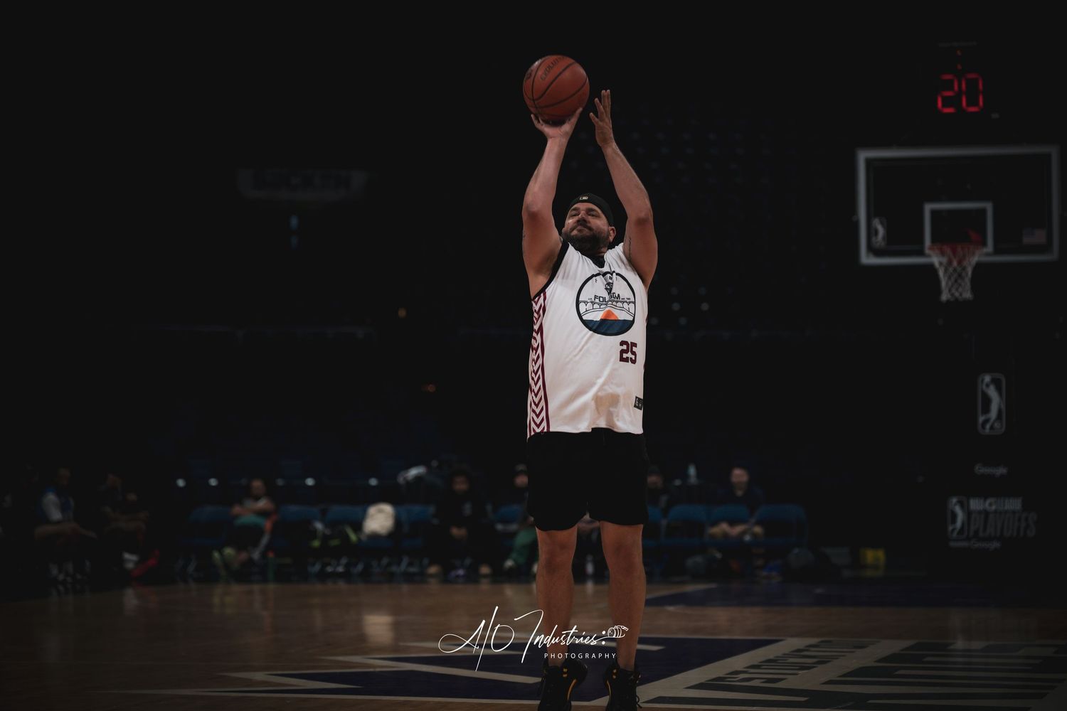 Basketball player in white jersey shoots the ball during an indoor game under dramatic arena lighting.