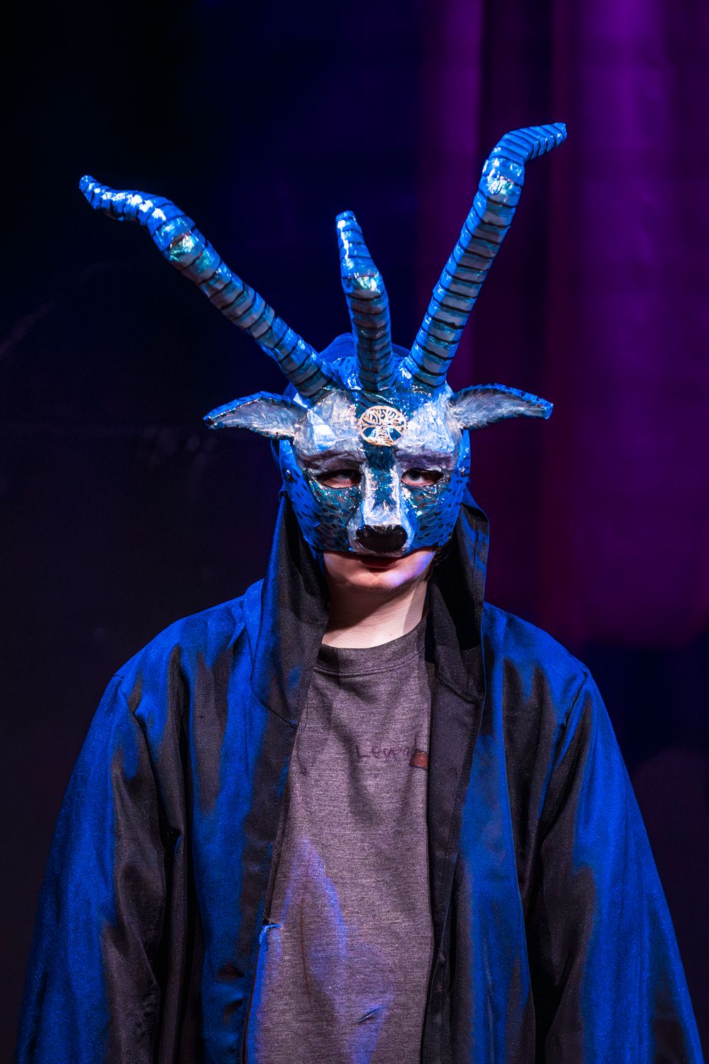 Person wearing a blue horned demon mask and dark cloak under dramatic stage lighting.