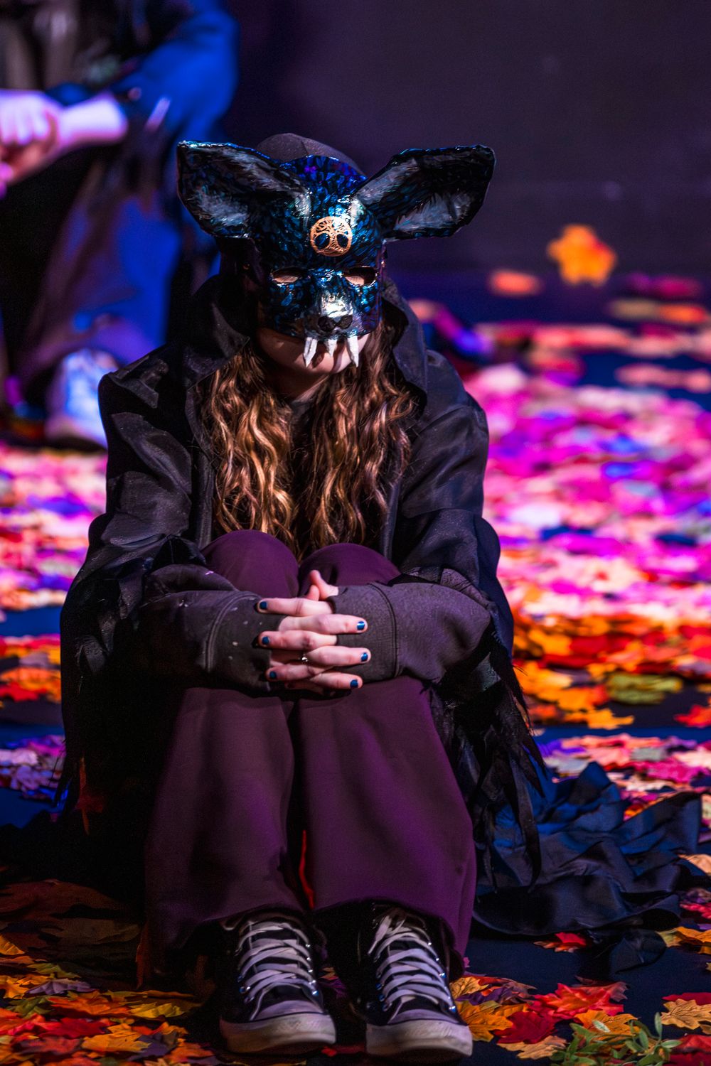Person in sequined fox mask sitting among colorful autumn leaves on a dark stage floor.