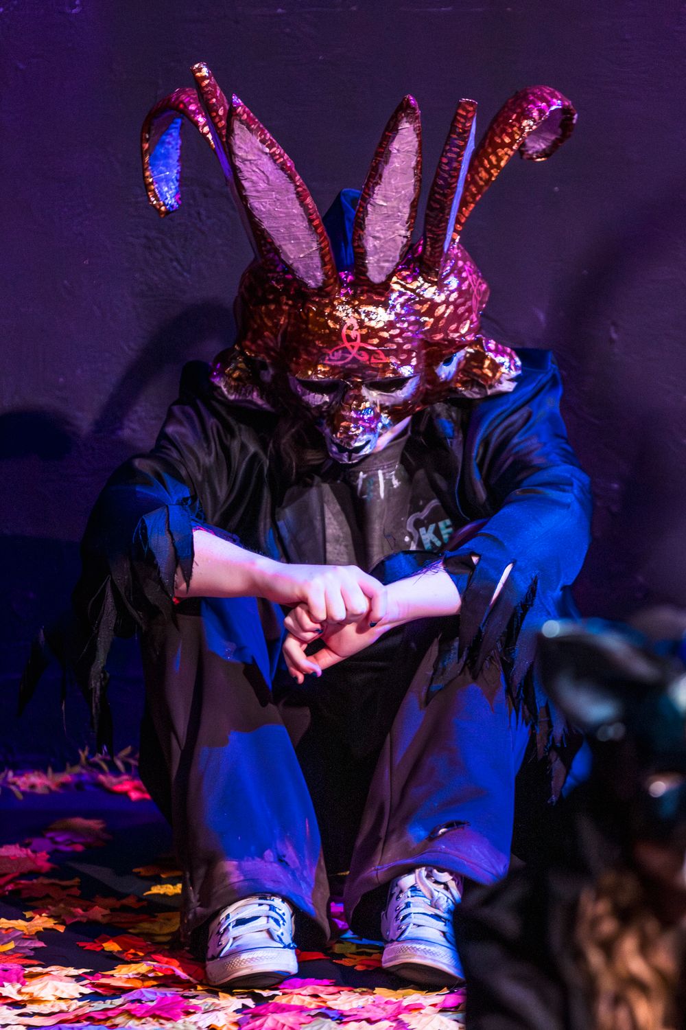 Person sitting on floor wearing shiny pink bunny mask and blue outfit under colorful party lights.