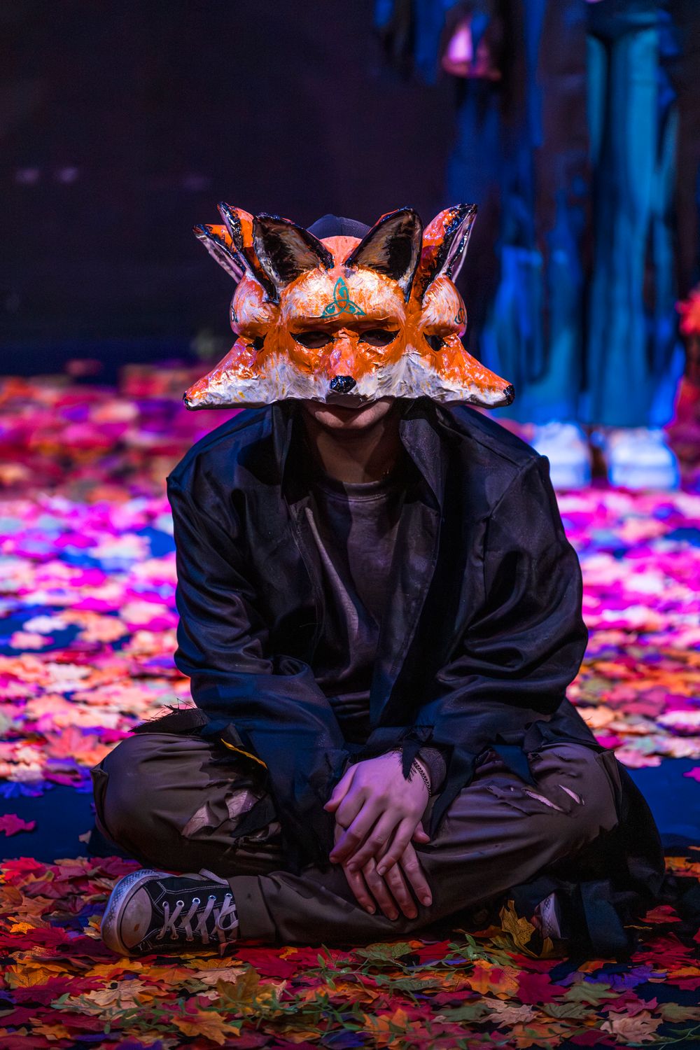 Person sitting cross-legged wearing an ornate fox mask surrounded by colorful autumn leaves on a stage.