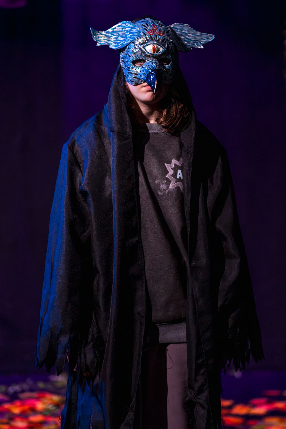 Person wearing blue dragon mask and black hooded cloak on stage.