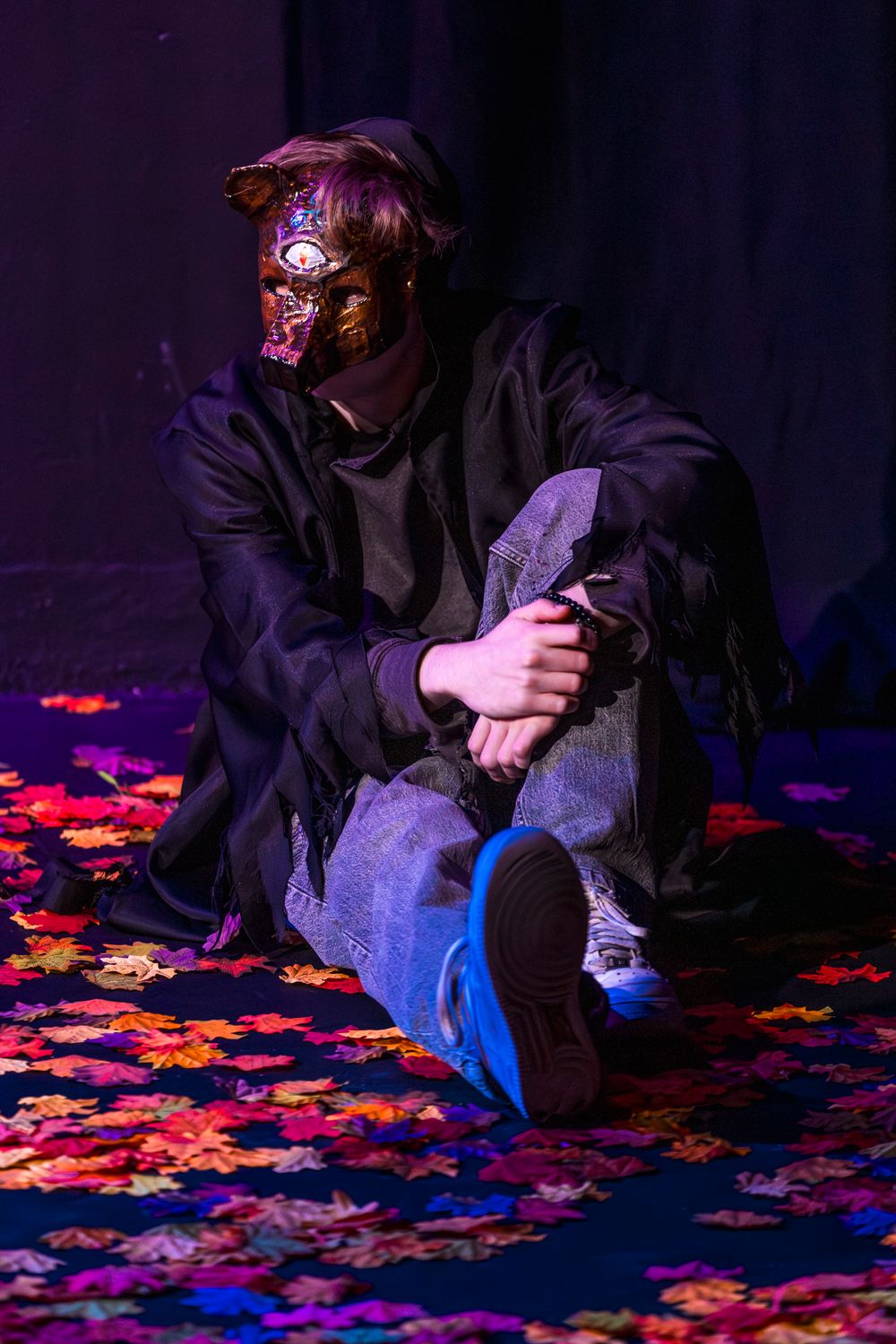 Person in gold mask sitting on colorful autumn leaves under purple stage lighting.