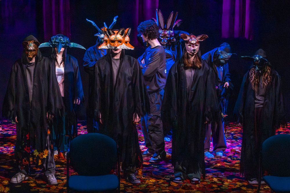 Performers in dark robes and colorful animal masks stand on a decorated stage under dramatic purple lighting.