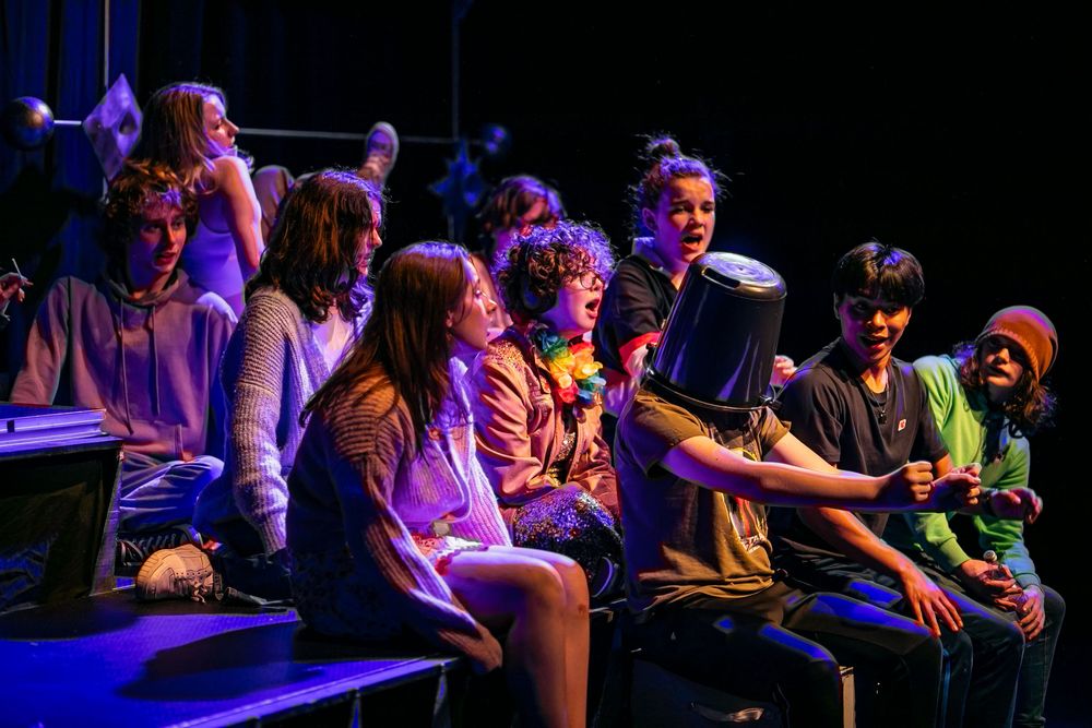 A group of people sit together watching a performance in a dark theater illuminated by purple stage lighting.