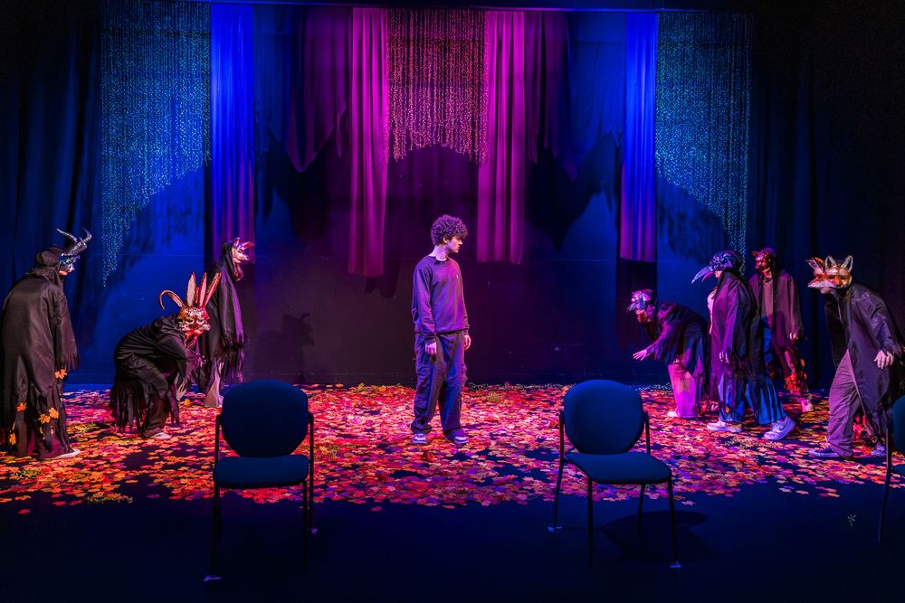 Theater performers in elaborate costumes surround a lone actor on a colorfully lit stage with scattered petals.