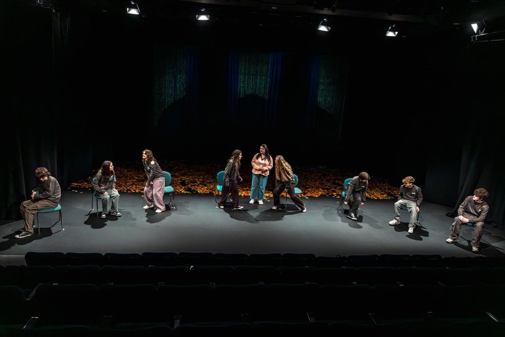 Theater performers on a dimly lit stage with floral backdrop, some seated and some standing during a dramatic scene.