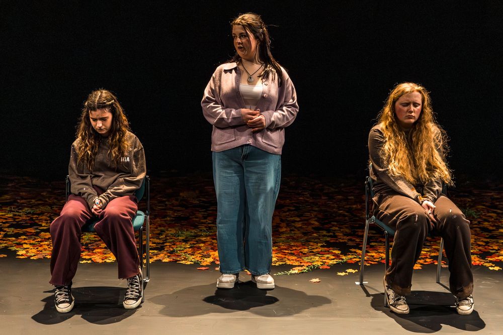 Three women perform on a dark stage with autumn leaf backdrop, two seated and one standing center.