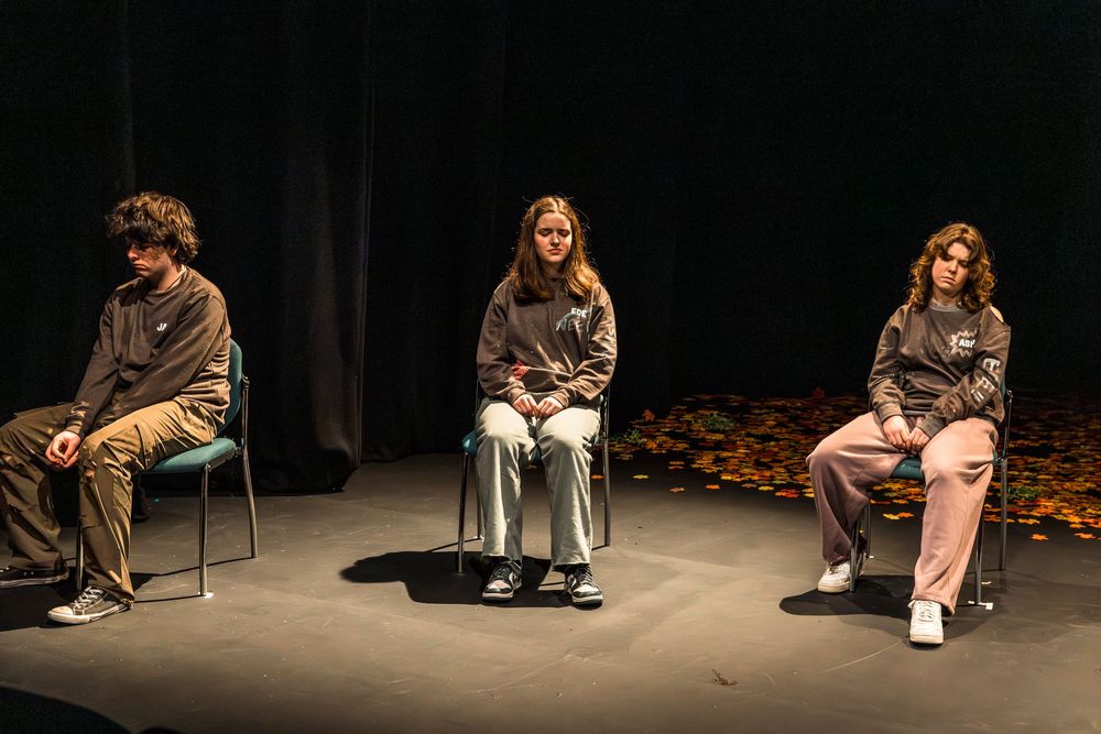 Three teens in matching gray hoodies sit on chairs on a dark theater stage with autumn leaves scattered on the floor.