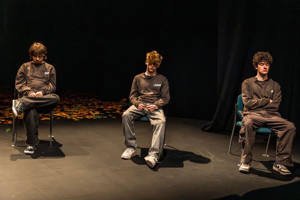 Three young men in gray hoodies sit on stools on a dark theater stage during a dramatic performance.
