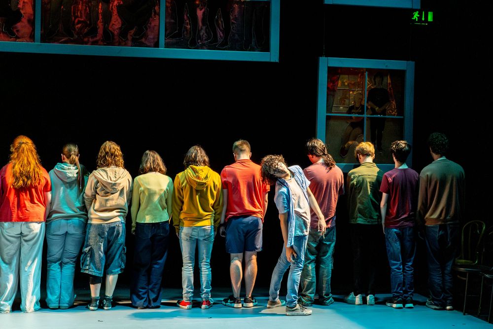 A line of people wearing colorful shirts and jeans stand with their backs turned to the camera on a stage.