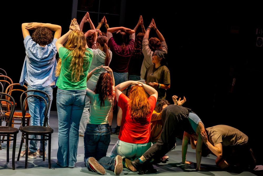 Performers on stage strike dramatic poses during a theatrical production with wooden chairs as props.
