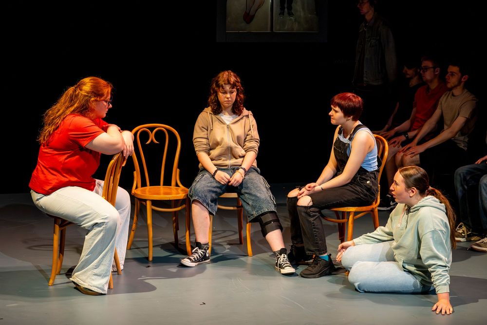 A group of performers sit in chairs arranged in a circle during what appears to be a theater workshop or rehearsal.