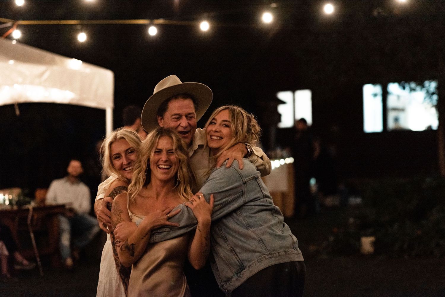 A group of people laugh together while celebrating outdoors under string lights at a nighttime gathering.