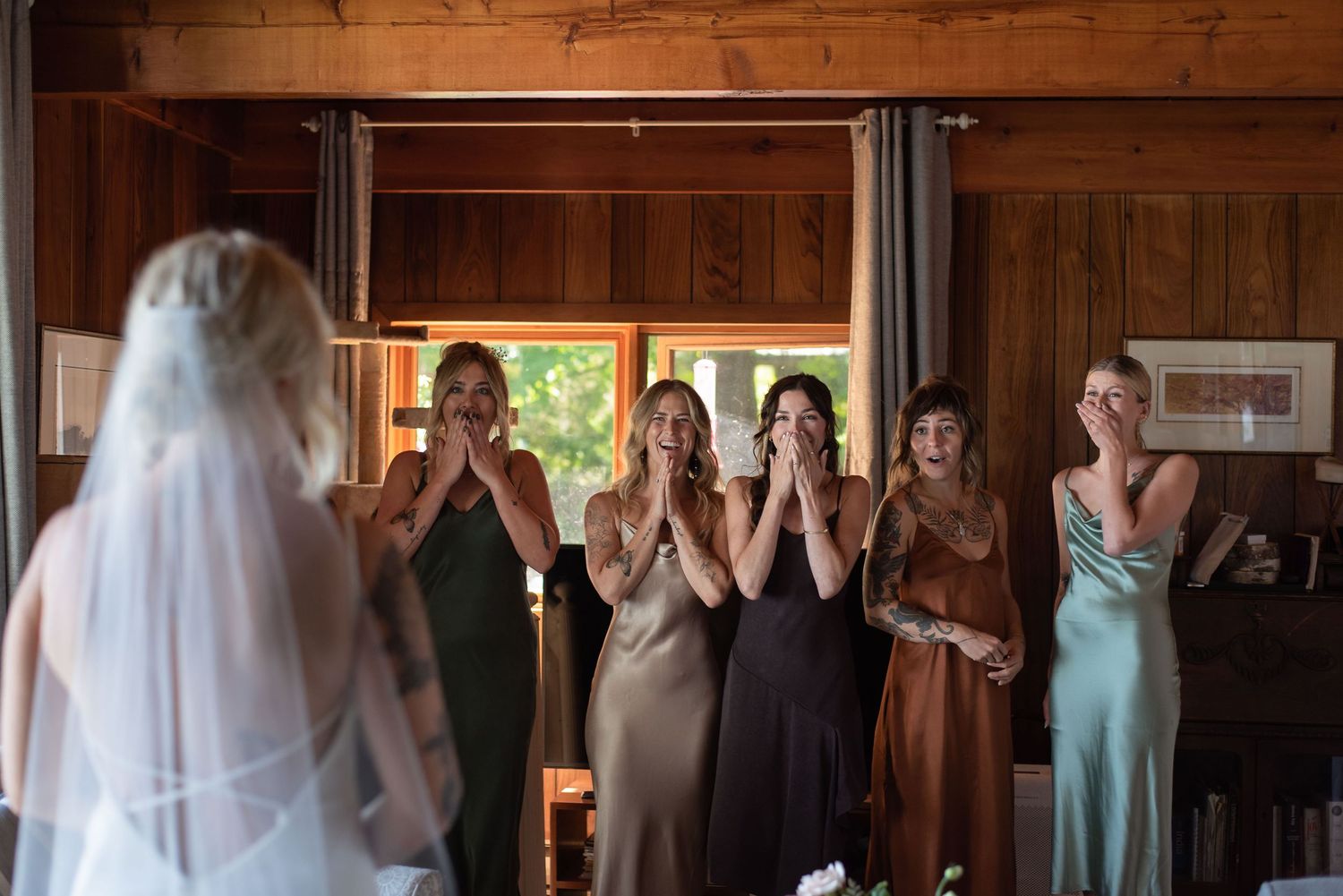 Bridesmaids react emotionally with hands covering mouths as bride in white veil approaches at indoor wedding venue.