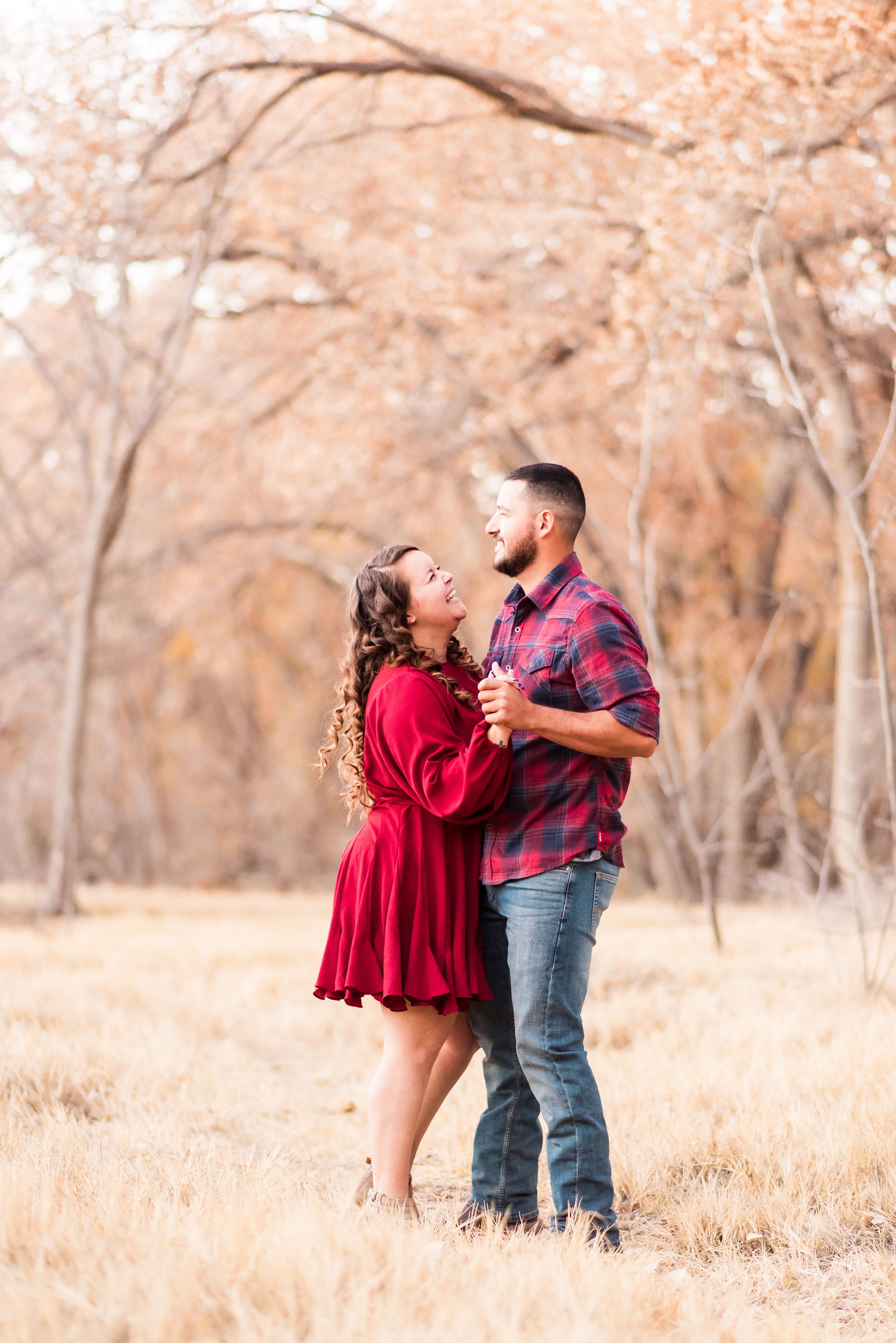 A couple dances together in a field of golden grass during autumn with bare trees in the background.
