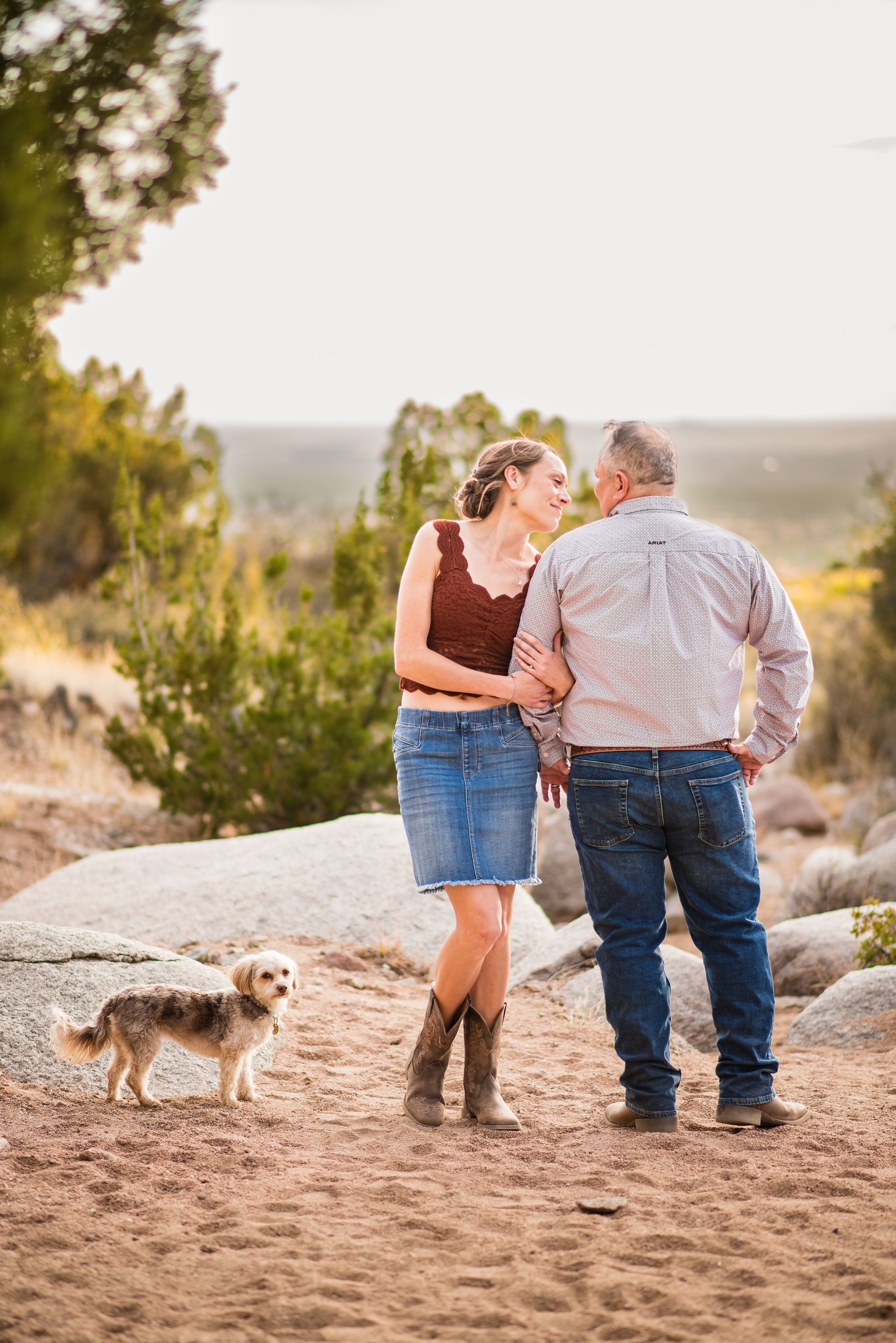 Couple in western attire with a small dog on a sandy trail surrounded by lush greenery during golden hour.