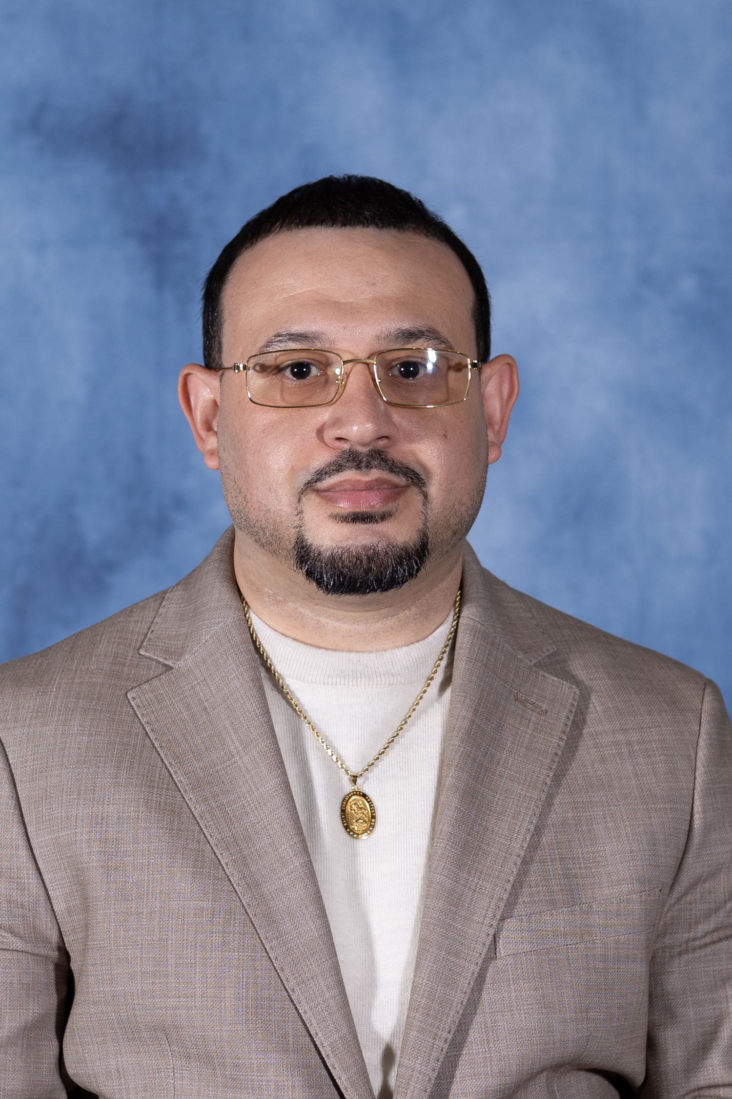 Professional headshot portrait against blue backdrop showing person in tan suit jacket, glasses and white shirt.