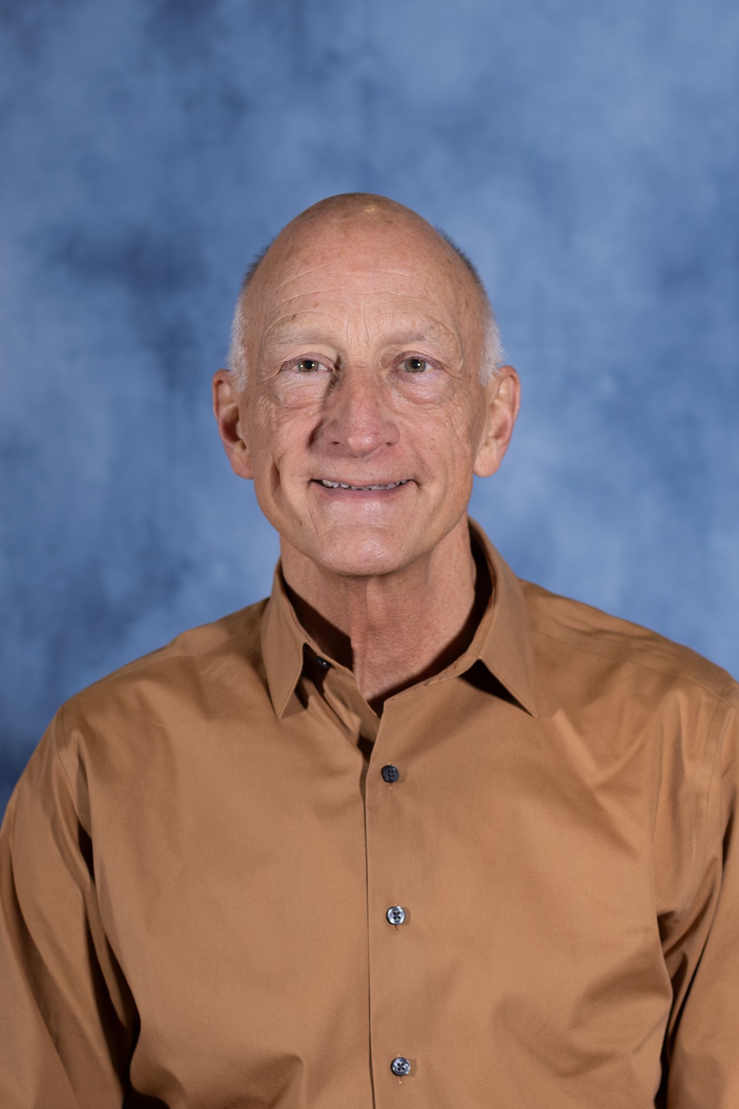 Professional headshot of a mature person in a brown collared shirt against a mottled blue background.