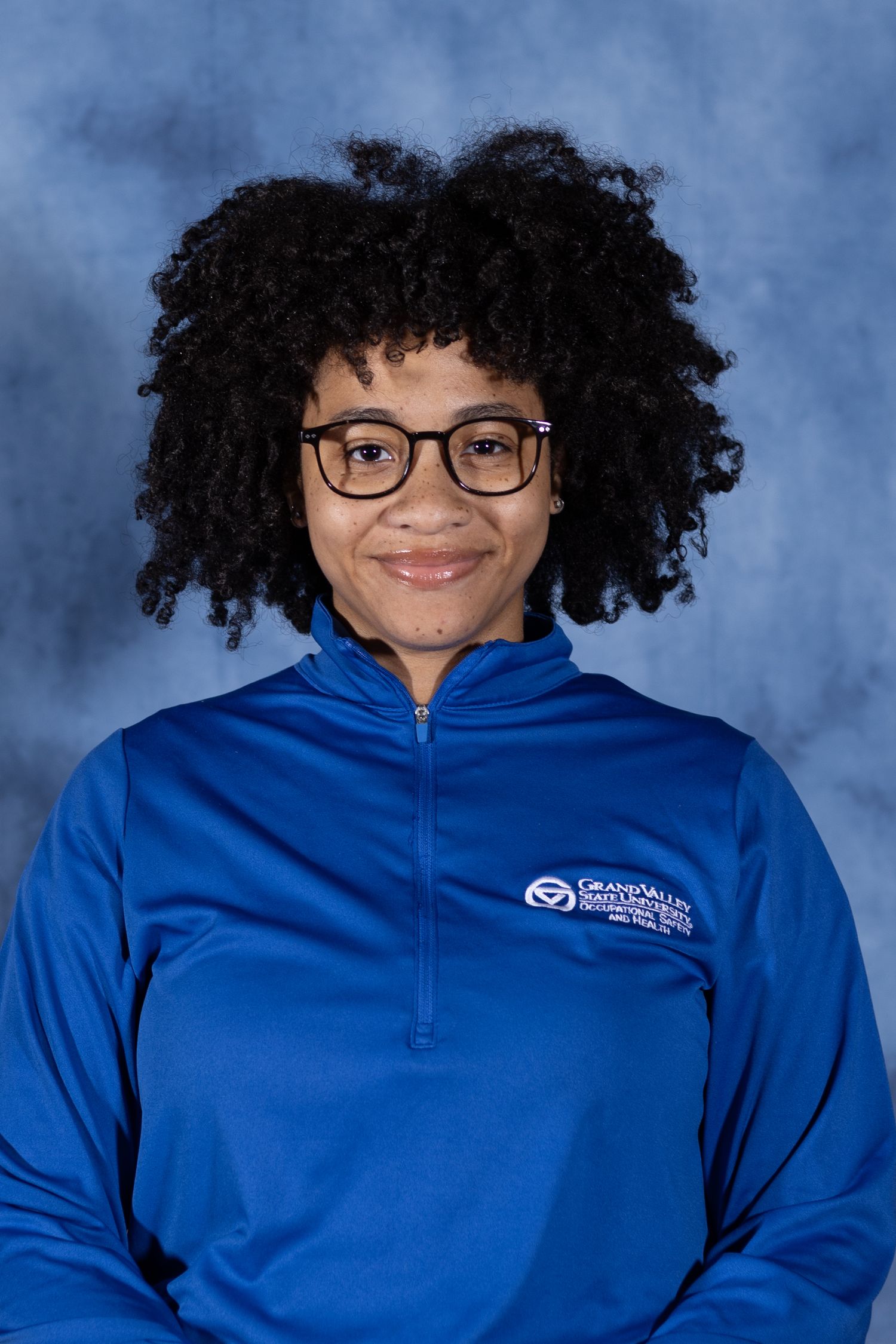 Professional portrait of someone in glasses and a blue quarter-zip pullover with curly natural hair against a blue backdrop.