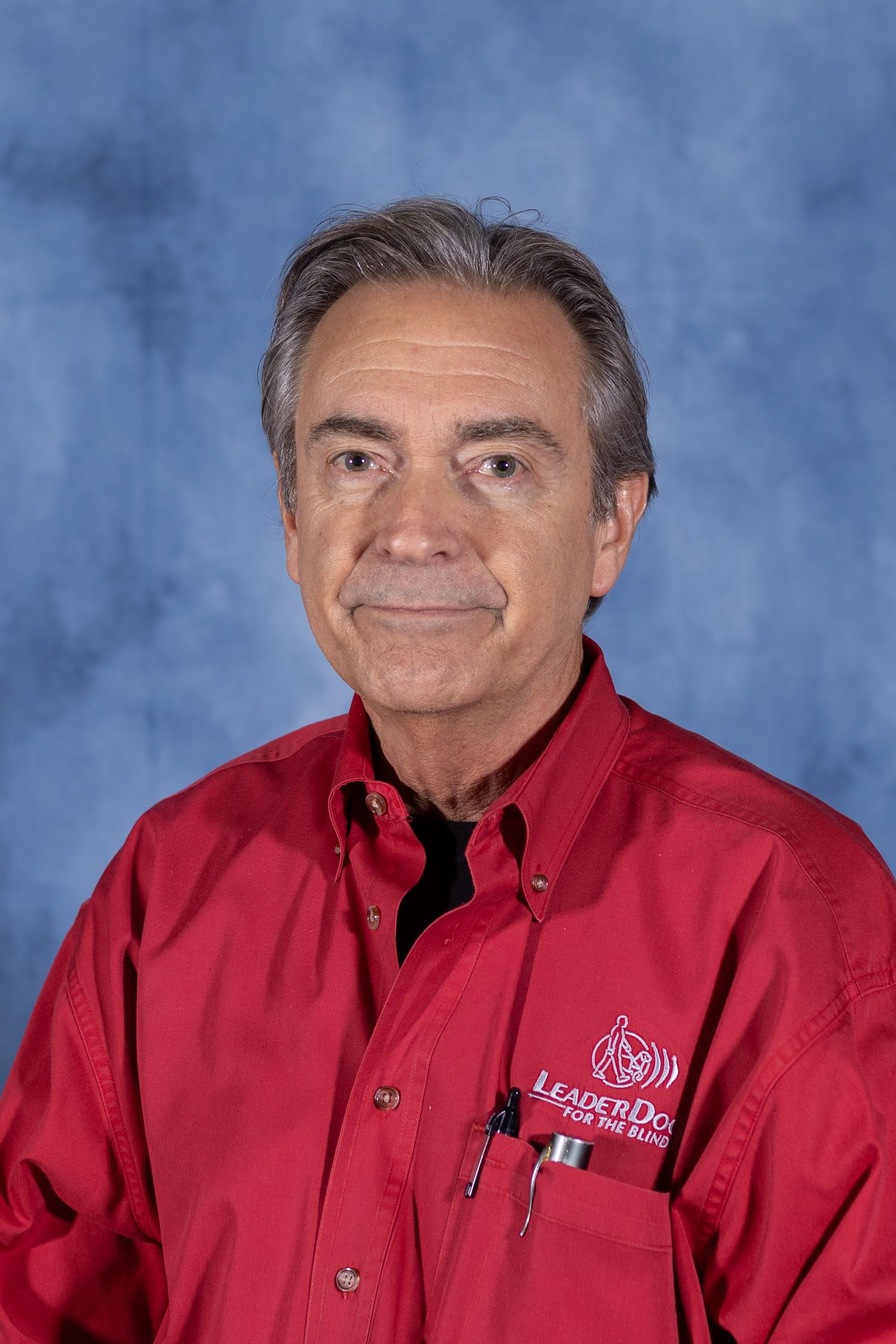 Professional headshot portrait of person in red button-down shirt with embroidered logo against blue background.