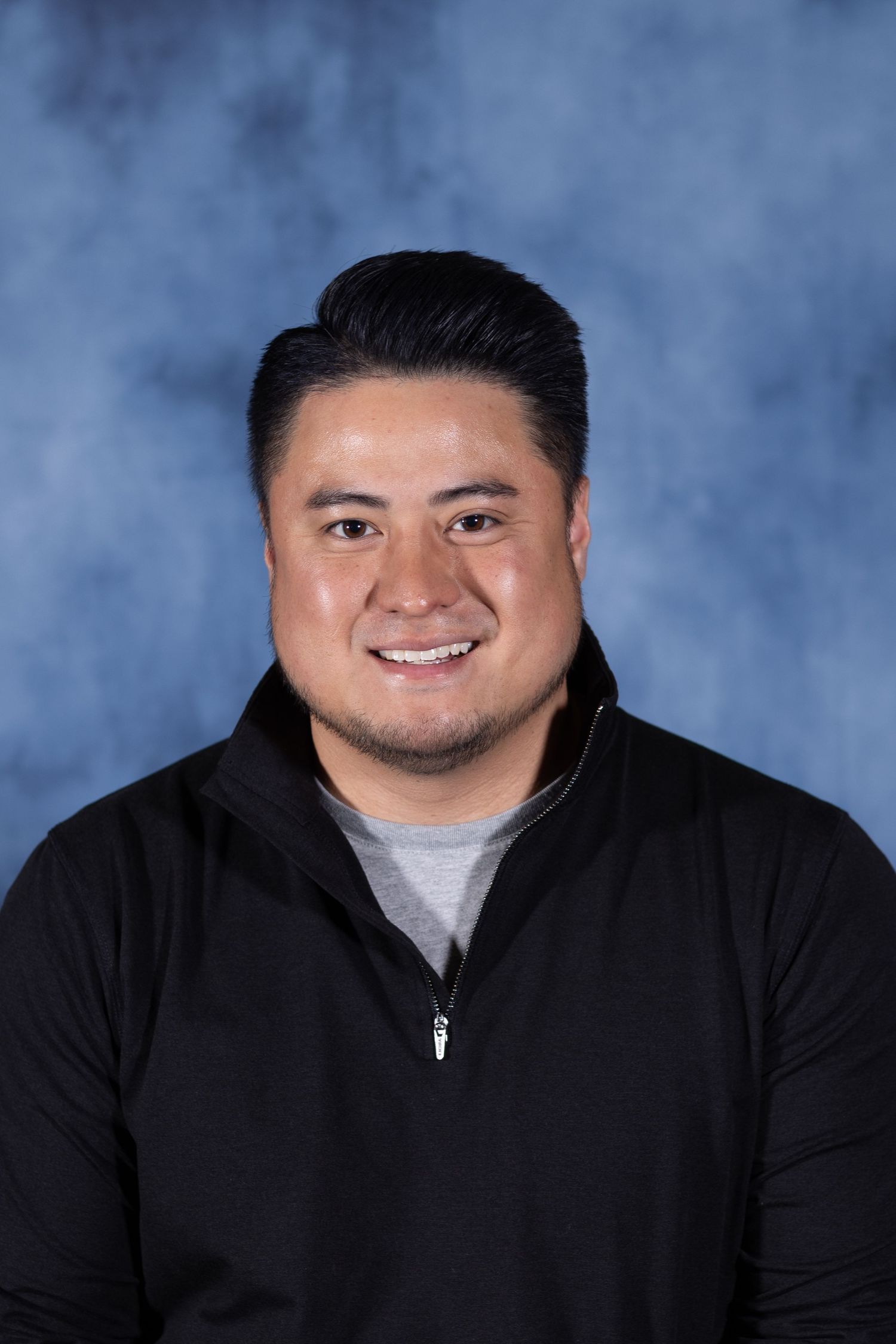 Professional headshot photo of someone wearing a black zip-up jacket and white t-shirt against a blue backdrop.