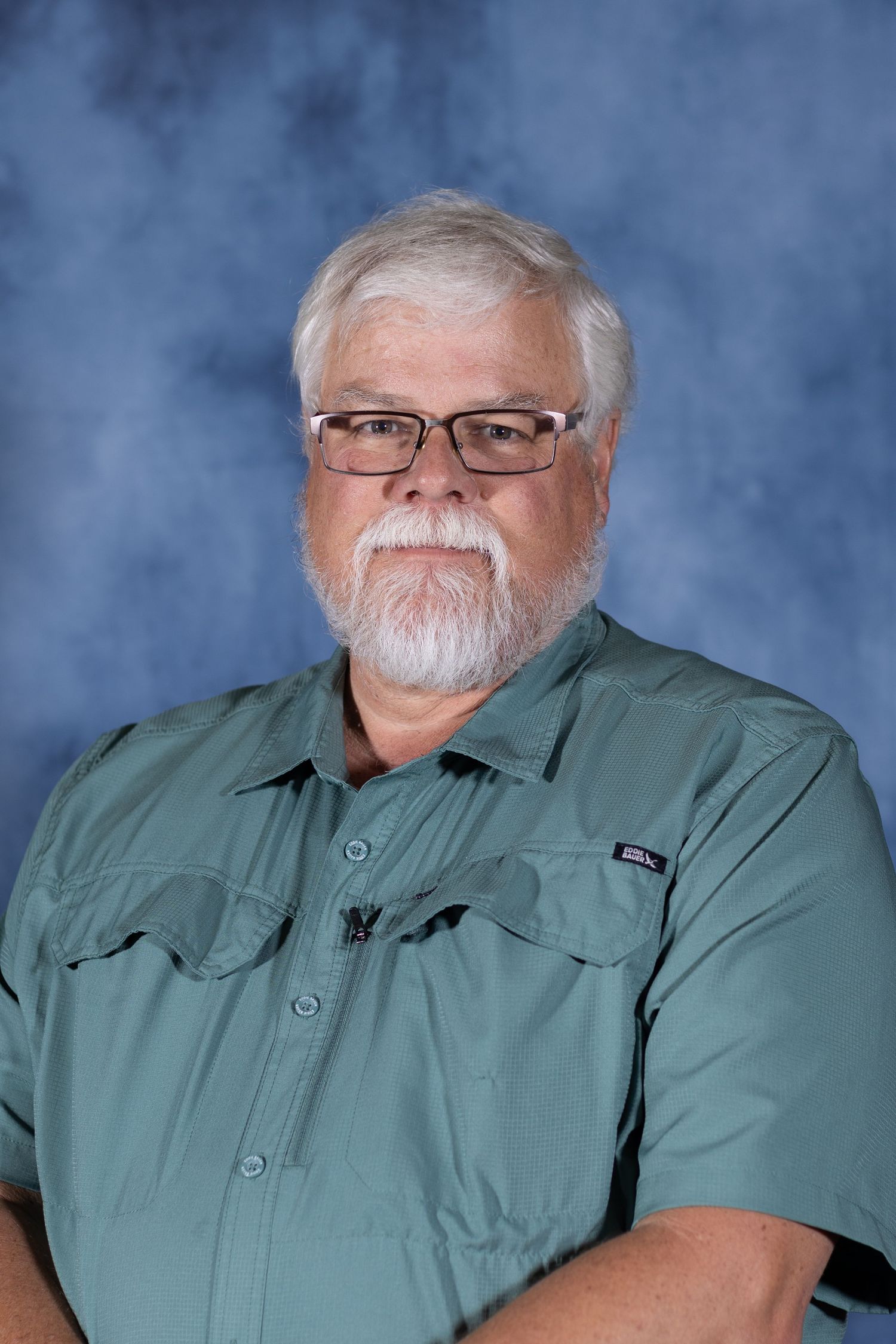 Professional portrait of an individual with white hair and beard wearing glasses and a sage green button-up shirt against blue backdrop.