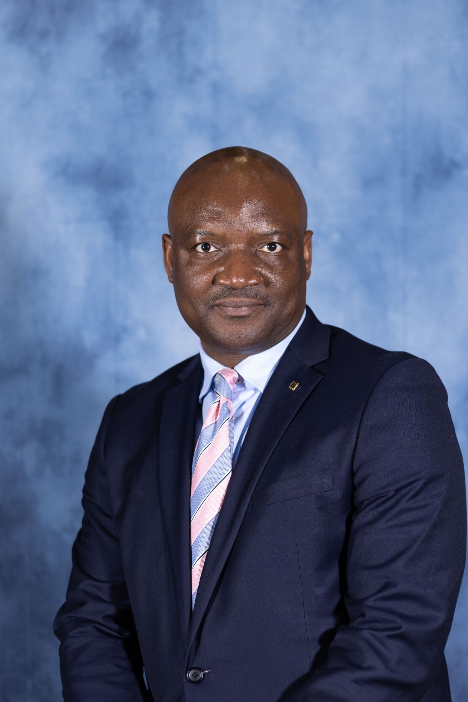 Professional headshot portrait of a person wearing a navy suit with pink striped tie against a blue backdrop.