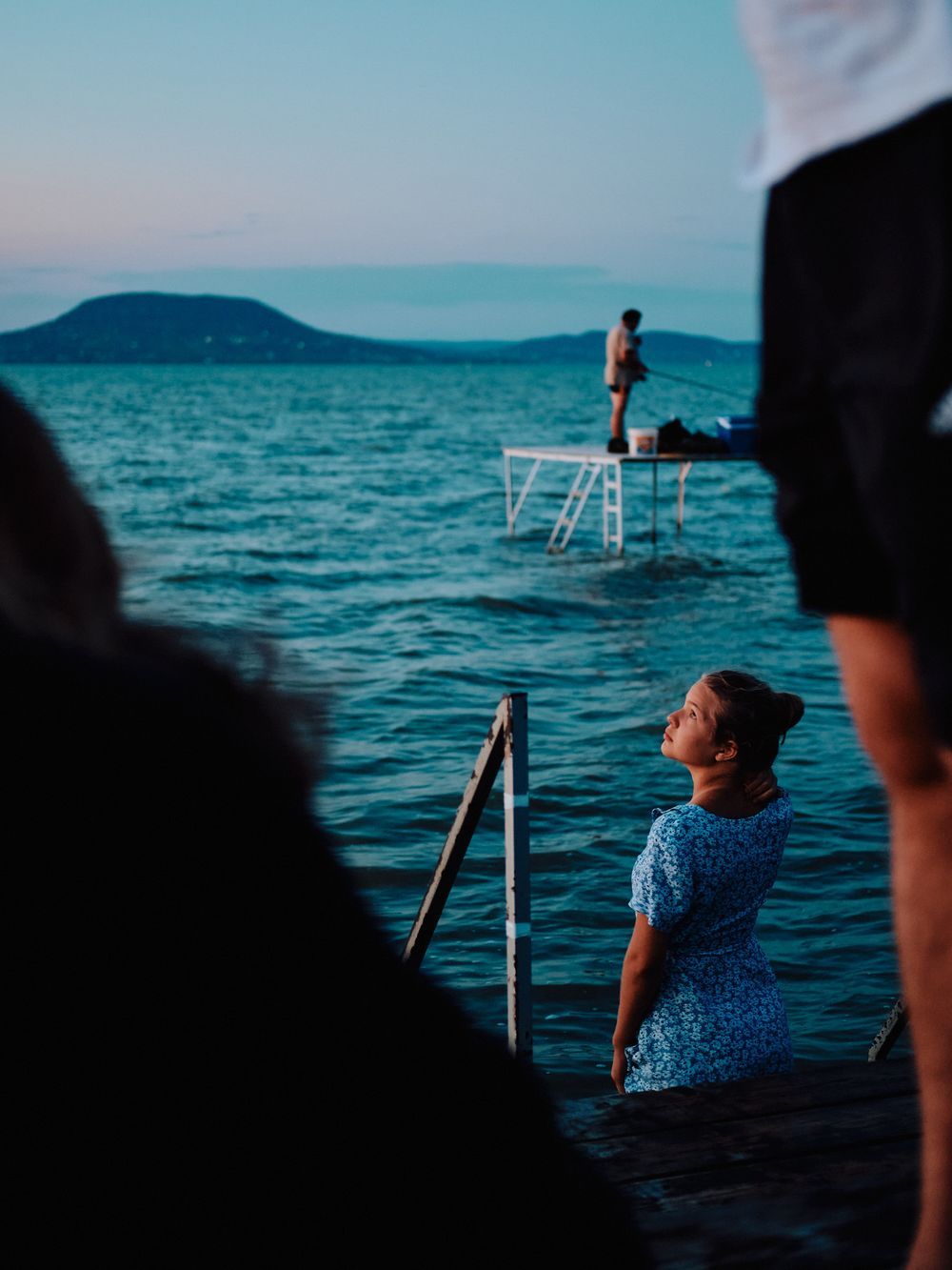 Spectators watch someone standing on a diving platform over Lake Balaton at dusk with mountains in the distance.