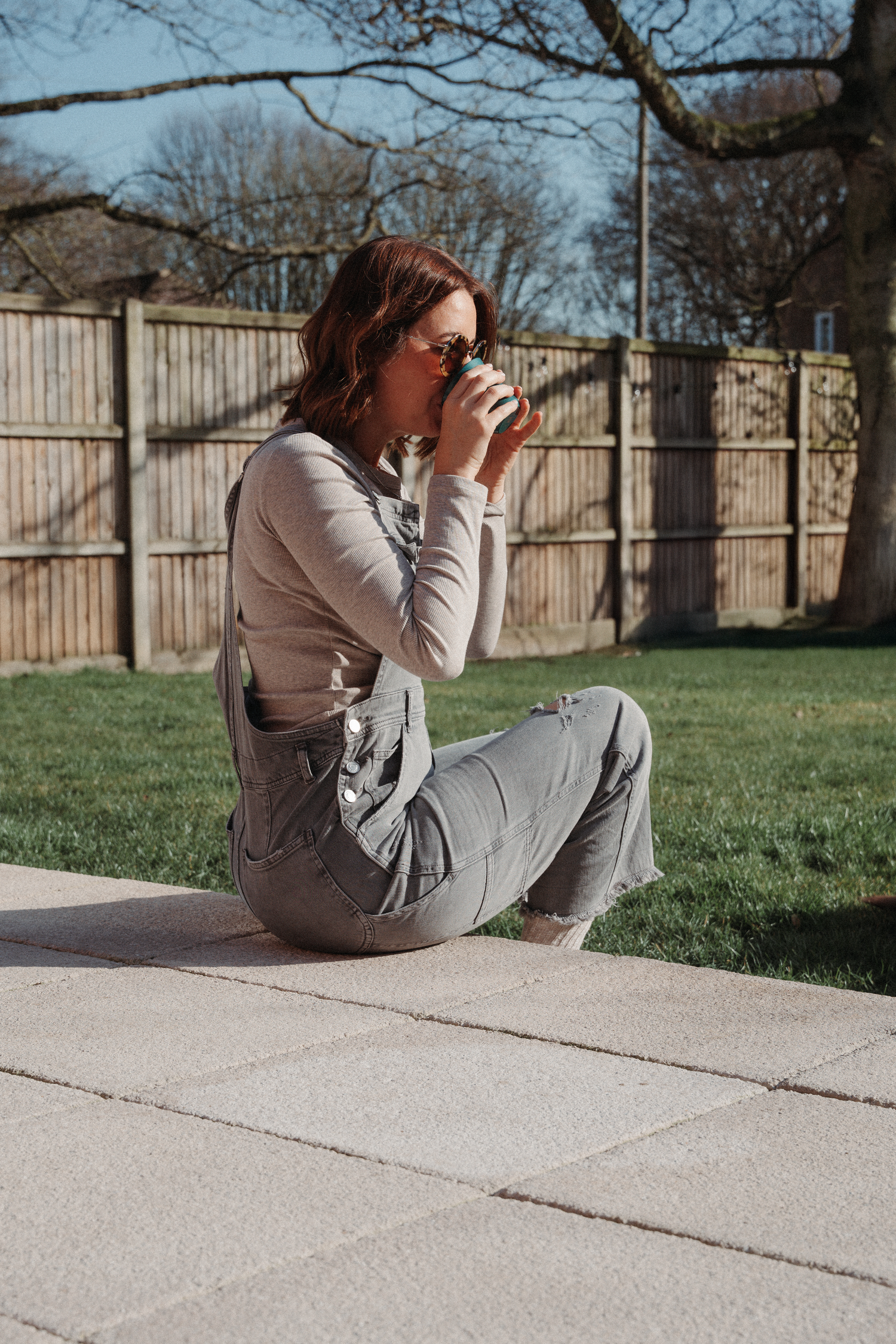 Person sipping a drink while sitting on a concrete patio in a backyard with a wooden fence.