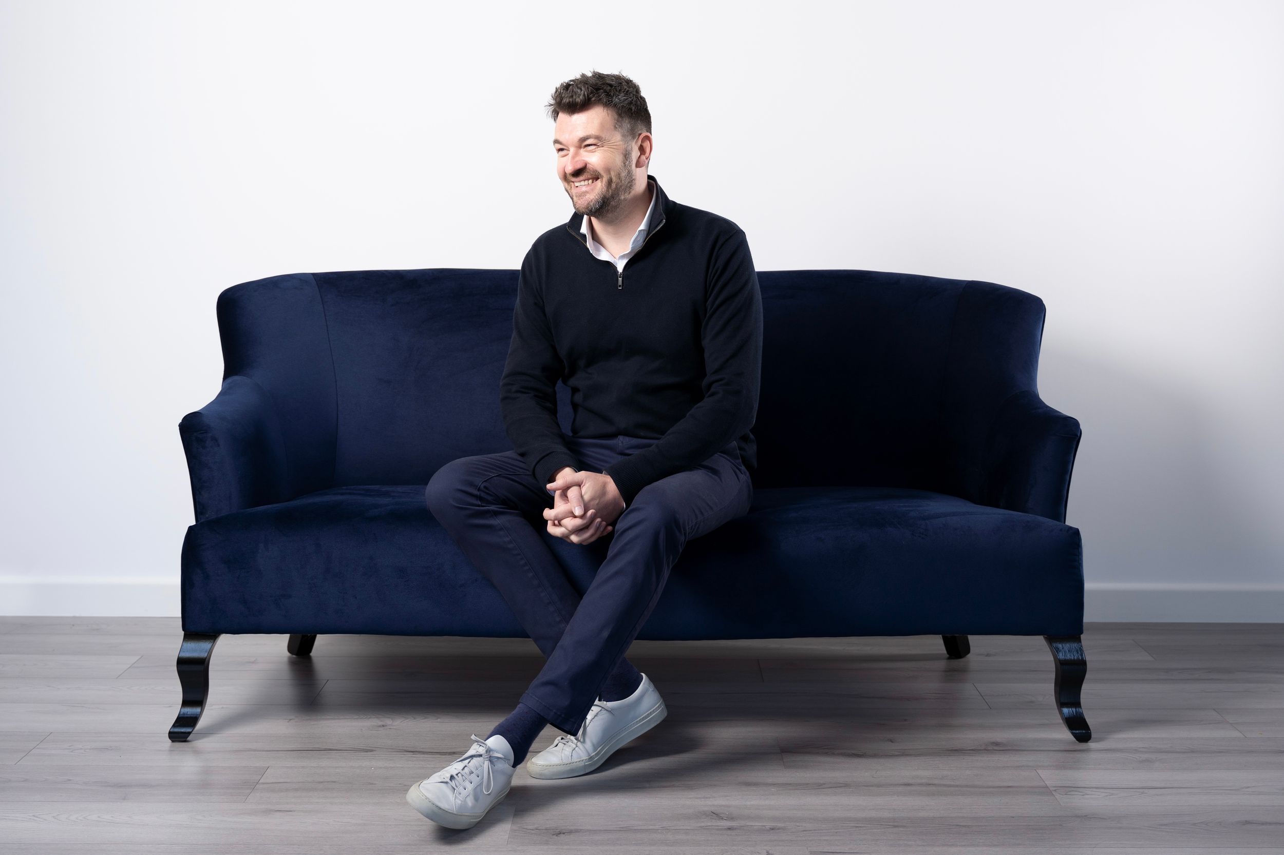 Male founder in dark clothing sits casually on a navy blue vintage sofa against a light grey wall on. personal branding photoshoot in London
