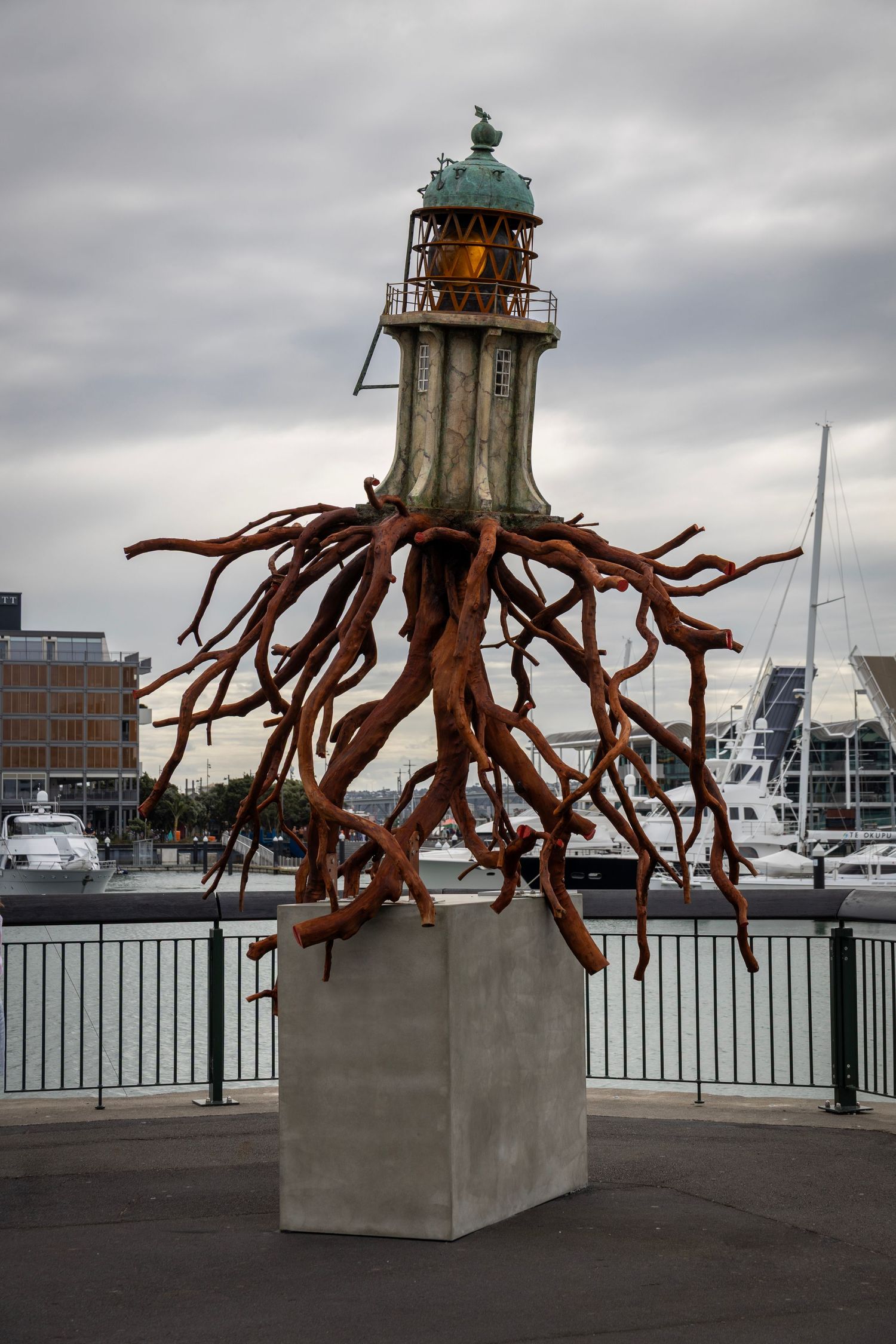 Sculpture of a lighthouse with sprawling tree roots, mounted on a concrete plinth in a waterfront urban setting with boats nearby.