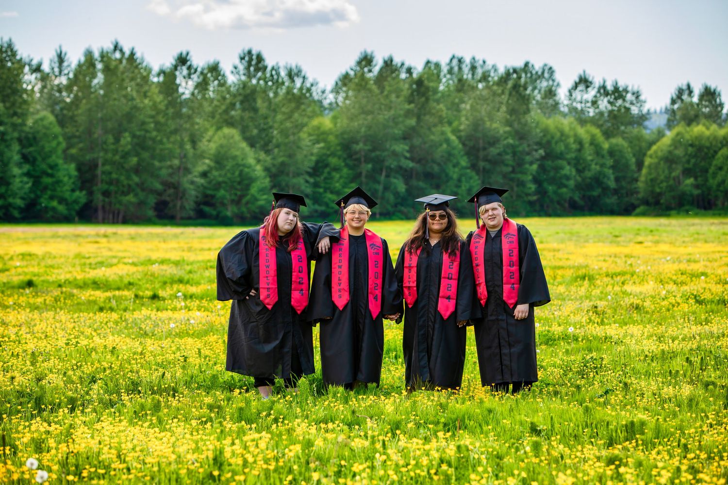 Four graduates in black caps and gowns with red stoles stand together in a vibrant yellow wildflower meadow.