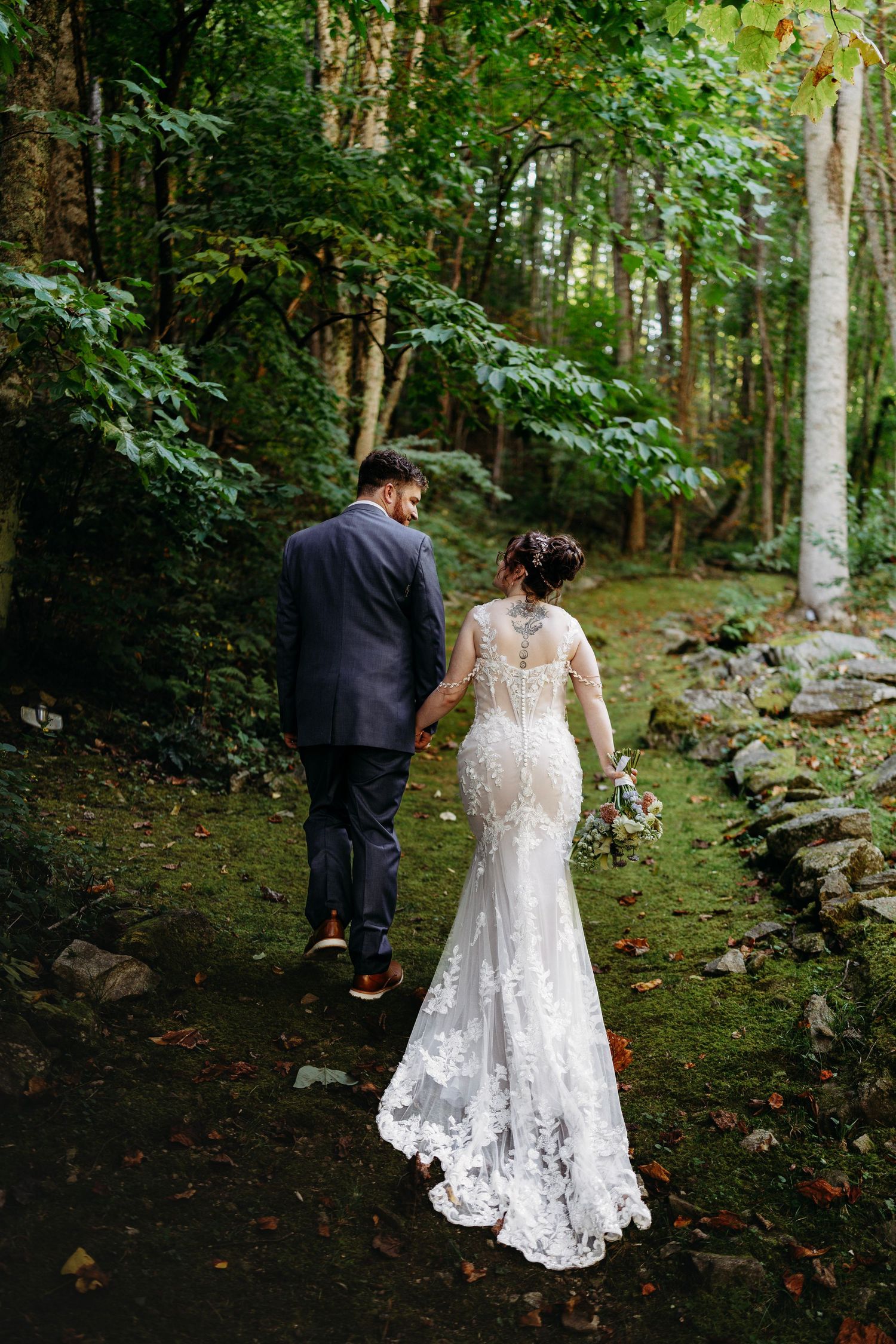 A bride in a lace mermaid gown and groom walk hand-in-hand along a lush forest path.