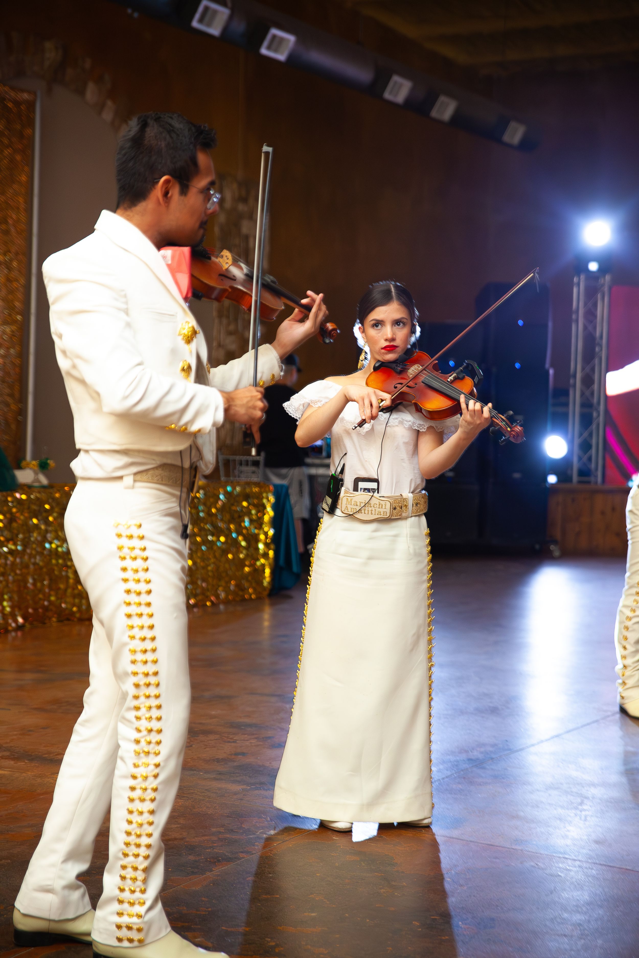 Musicians in traditional white mariachi outfits perform with violins on stage.